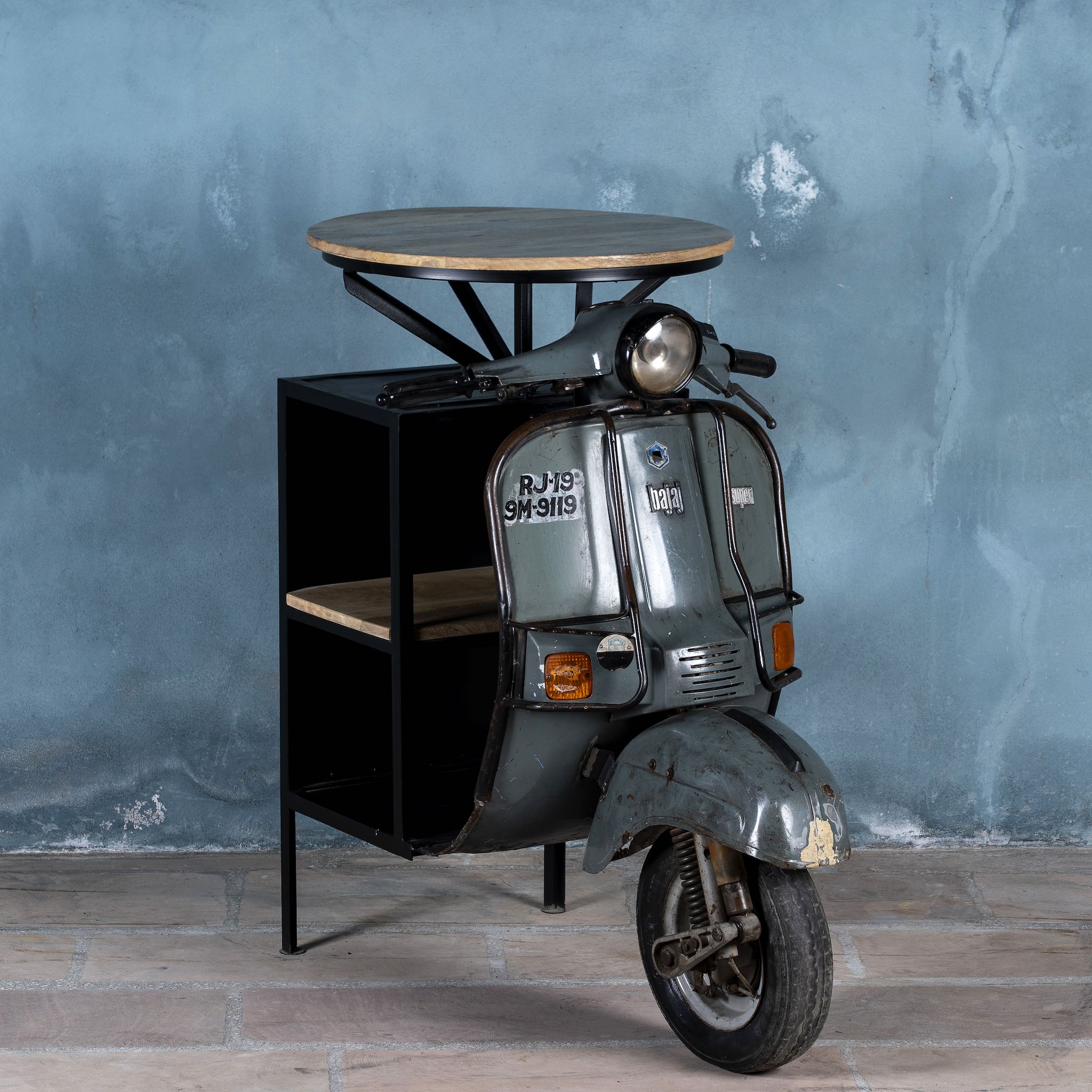 Side angle image of a short bar table crafted from the front half of an original Bajaj scooter, featuring a round wooden top, metal frame, and rustic industrial finish.