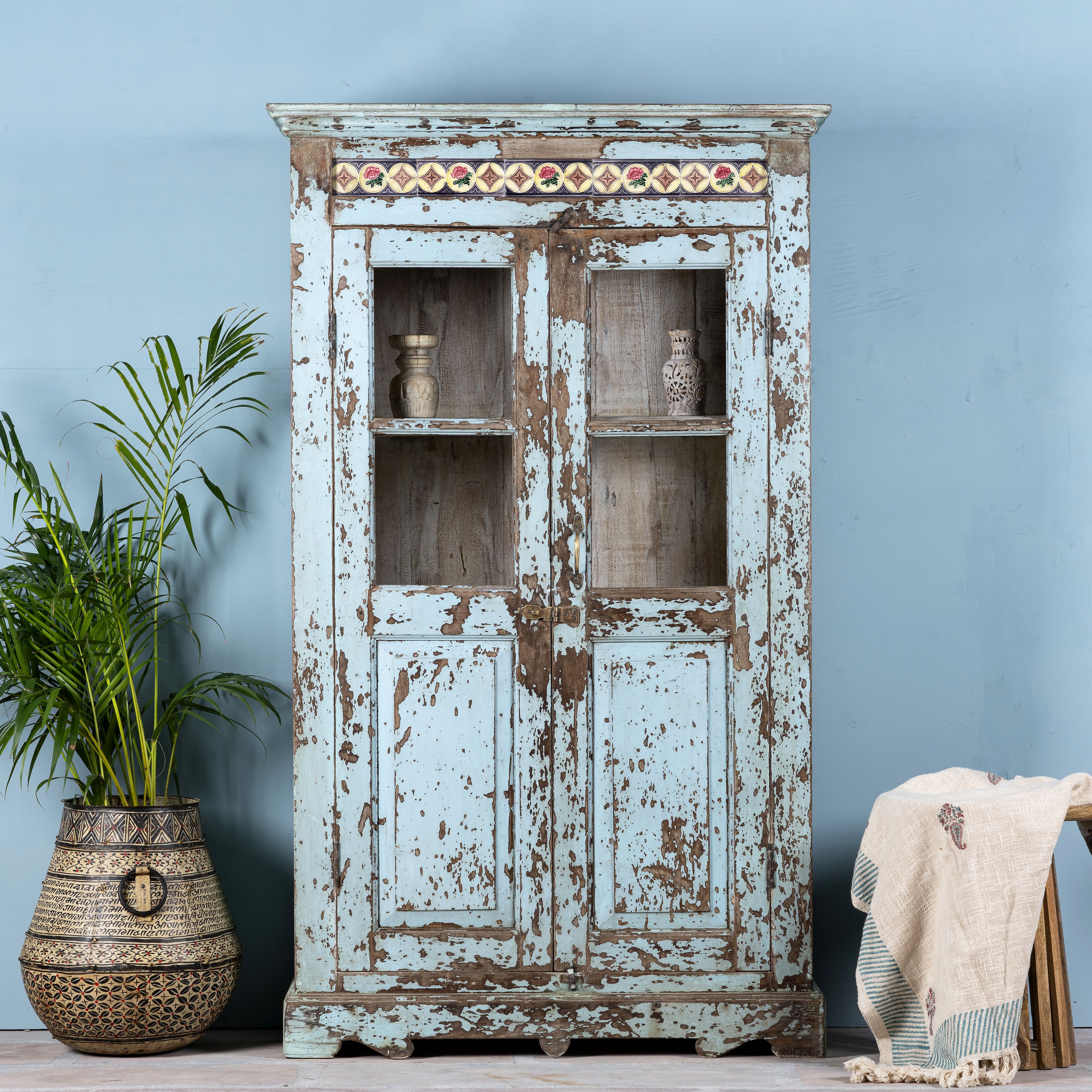 A light blue, antique teak wood showcase with ceramics, featuring two glass doors and multiple shelves, placed against a blue wall with a palm plant and a traditional textile behind it.