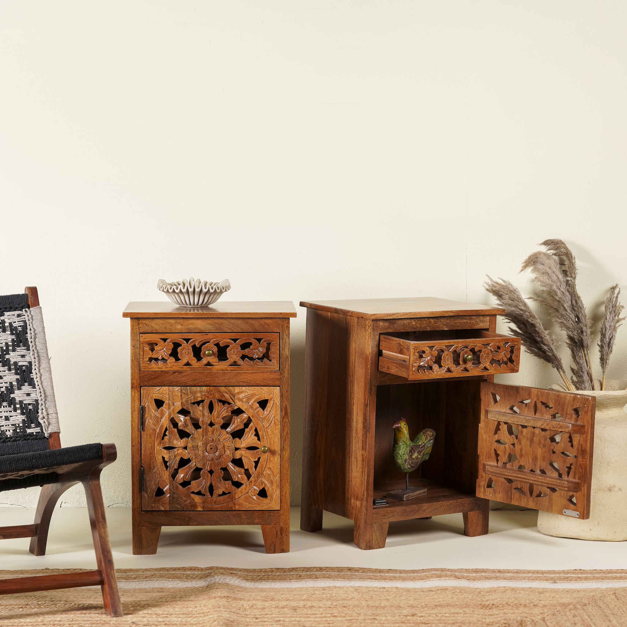 Side angle image with open door of a pair of mango wood night stands with hand carved floral and geometric detailing, warm teak finish; elegant hand carved furniture, 45 × 35 × 65 cm.