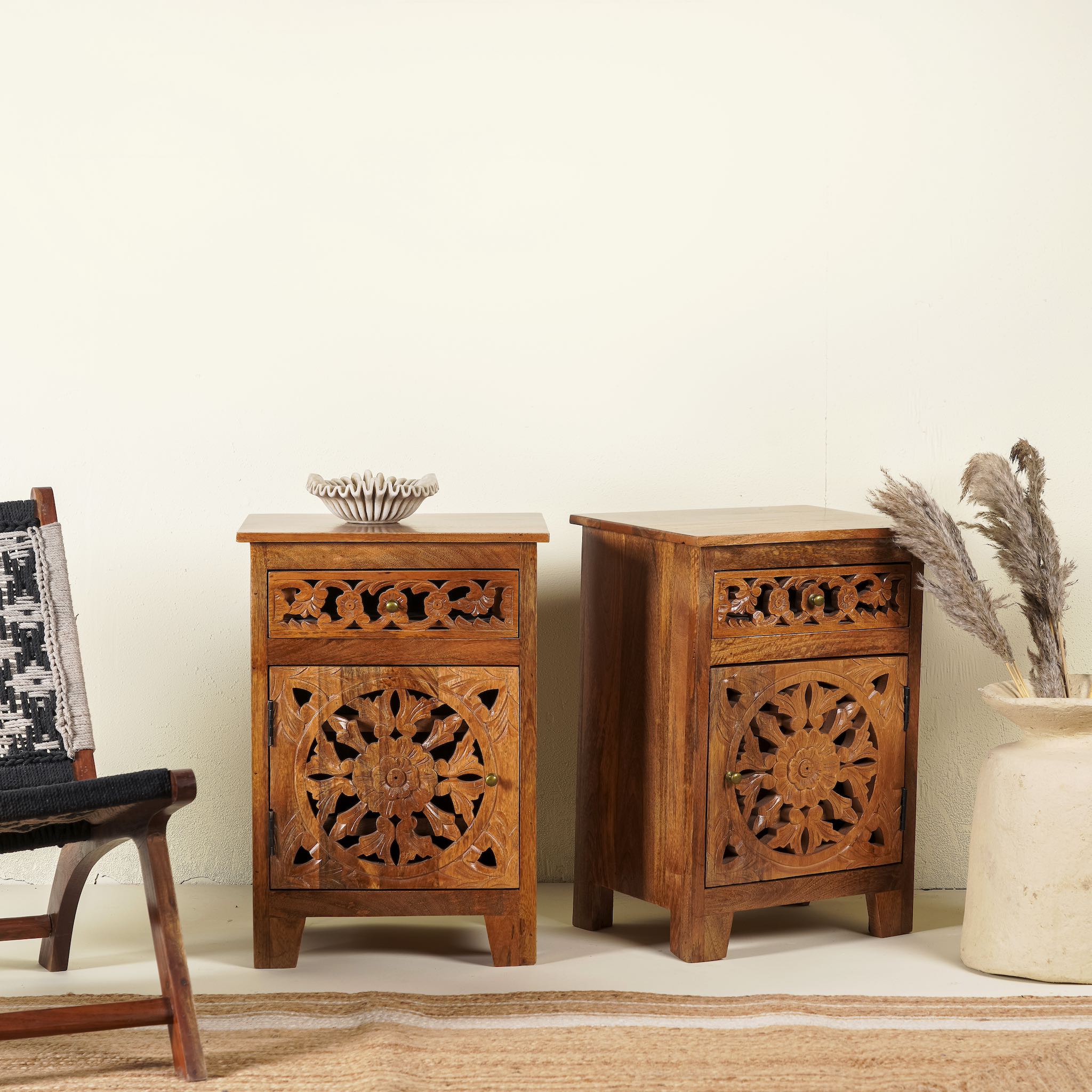 Side angle image of a pair of mango wood night stands with hand carved floral and geometric detailing, warm teak finish; elegant hand carved furniture, 45 × 35 × 65 cm.