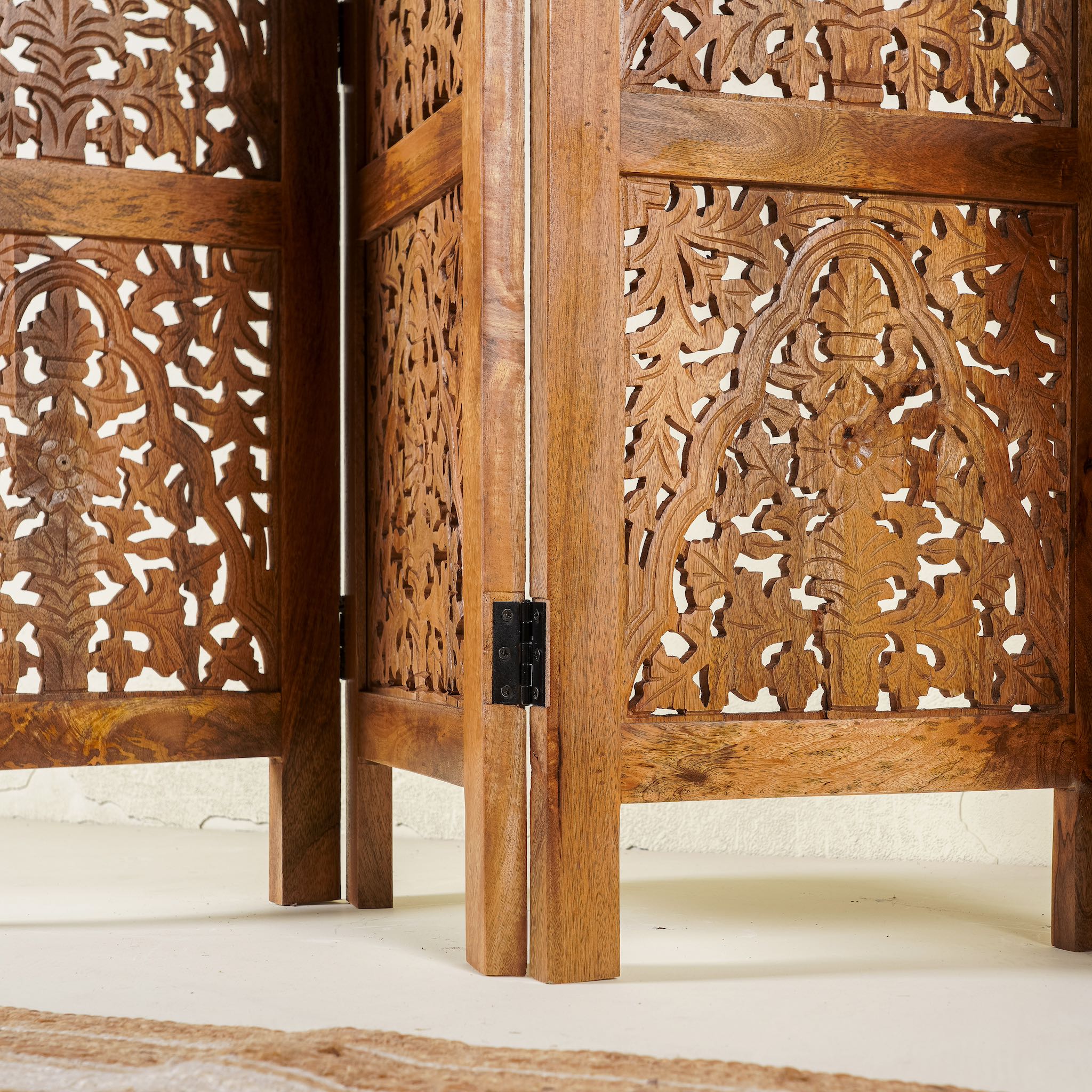 Close up image of a foldable room partition made from mango wood, featuring four panels with intricate hand-carved floral and lattice designs, finished in a warm natural tone and connected by black metal hinges for flexible positioning.