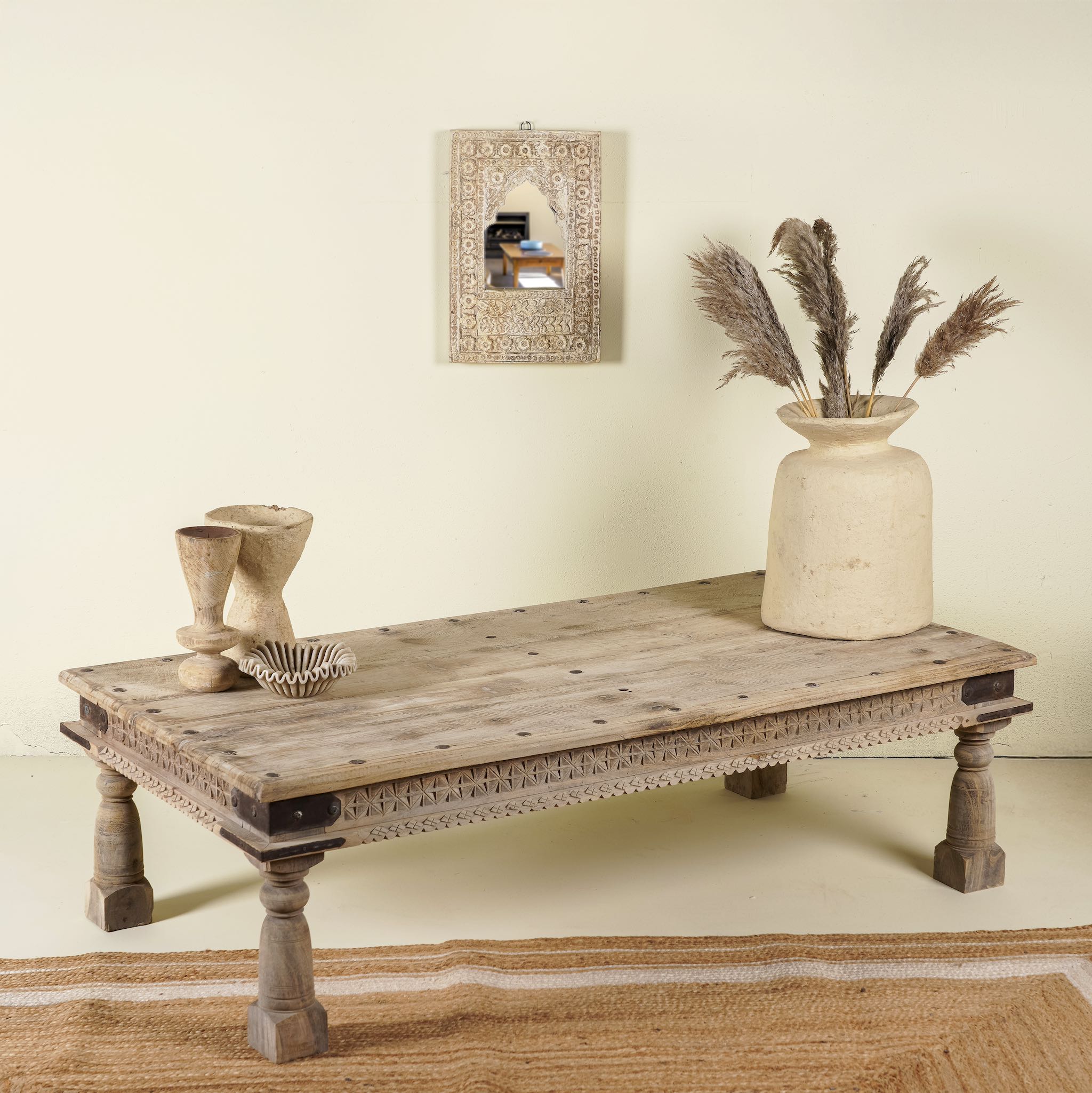 Rustic coffee table Amana made from old reclaimed teak wood, featuring a weathered finish, turned legs, decorative carved apron, and visible metal rivets, styled with earthy decor in a minimal interior setting.