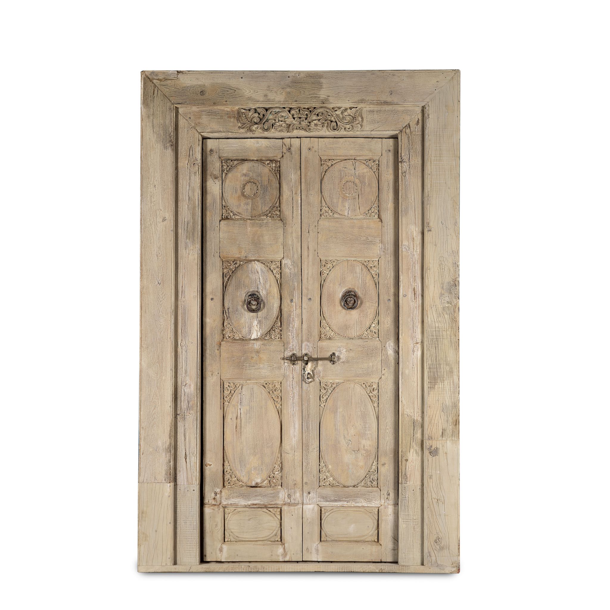 White background image of a antique Indian door Amaya made of reclaimed teak with brass fittings, hand-carved details, extended frame, usable as entrance or vintage wall panels, 161 × 20 × 250 cm.