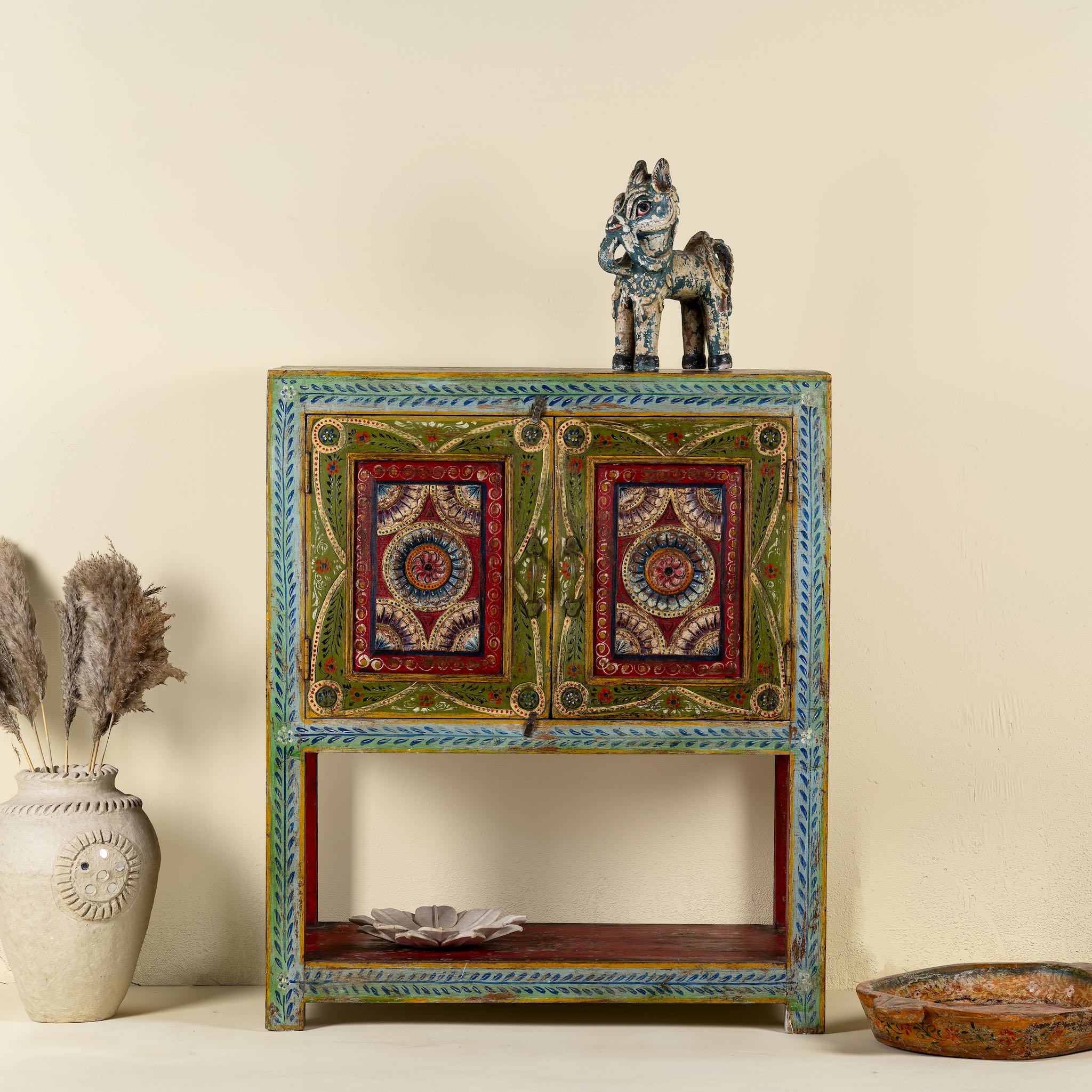 Front view image of a small hand-painted console made from reclaimed wood, featuring vibrant decorative motifs in red, green, blue, and gold on double doors, with intricate floral and geometric patterns and a lower open shelf for added display.