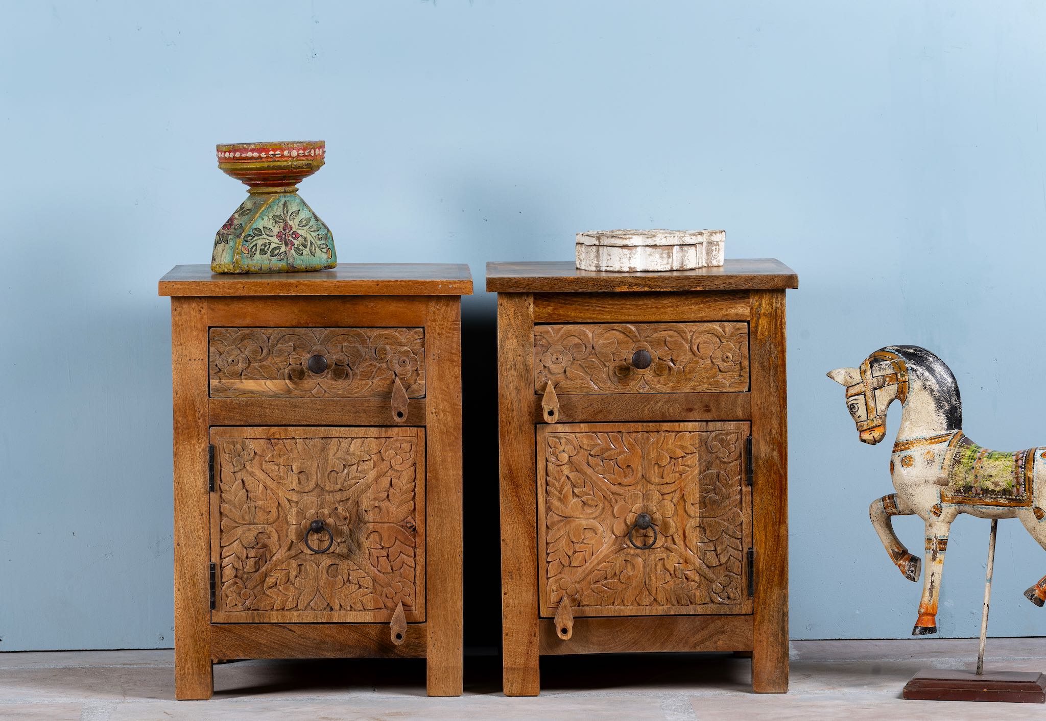 Two hand-carved mango wood bedside tables with floral patterns against a blue background.