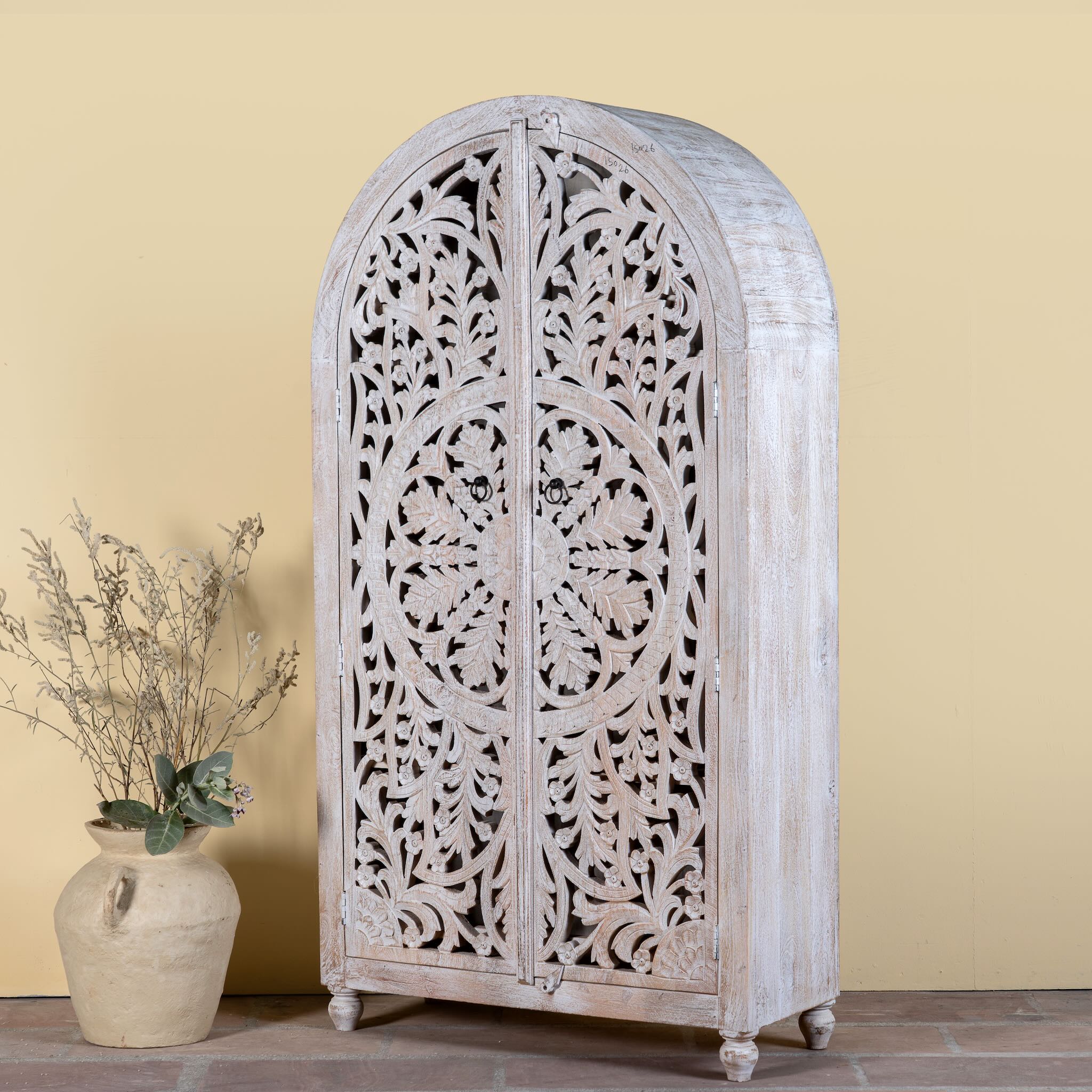 Side angle image of a Bhuvana arched mango wood wardrobe featuring a sculptural half-round design, intricate hand-carved floral detailing, and carved legs—an artisanal piece in the collection of arched wardrobes and mango wood wardrobes.