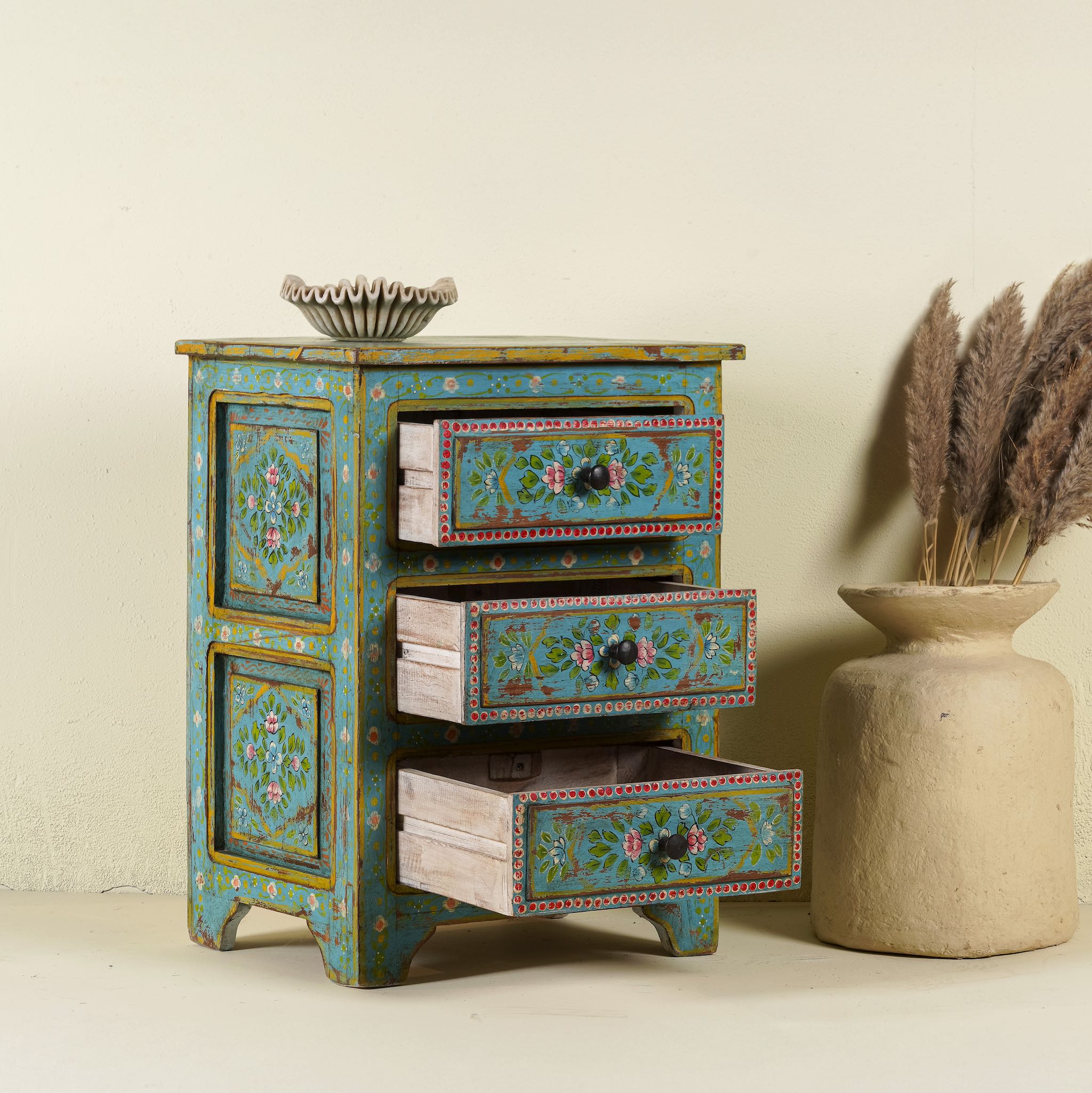 Open drawer image of a hand-painted reclaimed wood nightstand with vibrant colours and floral patterns, featuring three drawers, placed next to a vase with dried plants.
