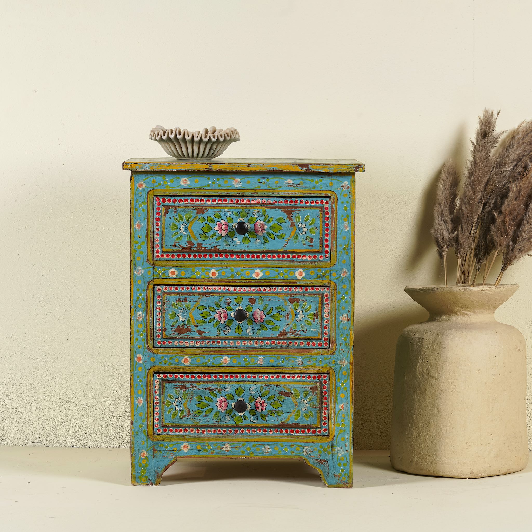 A hand-painted reclaimed wood nightstand with vibrant colours and floral patterns, featuring three drawers, placed next to a vase with dried plants.
