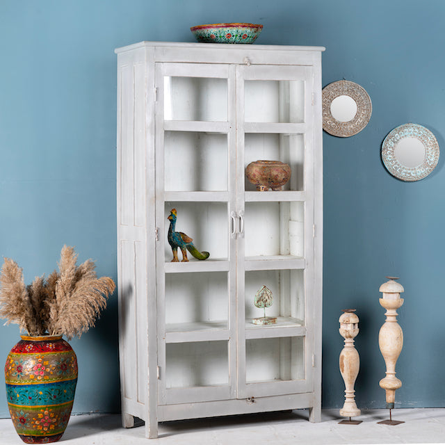 Side angle image of a shabby chic teak wood cabinet with glass doors and five compartments – sustainable living room furniture crafted from refurbished old teak with rustic white finish.