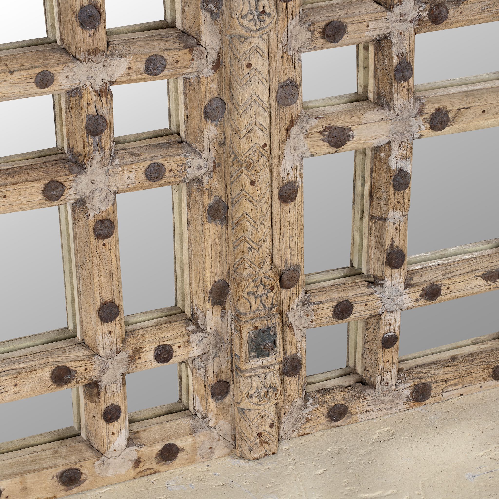 Close up of a statement wall mirror made from an old teak window frame with large square grid pattern, metal studs, and decorative center carving- a bold blend of rustic charm and historic craftsmanship.
