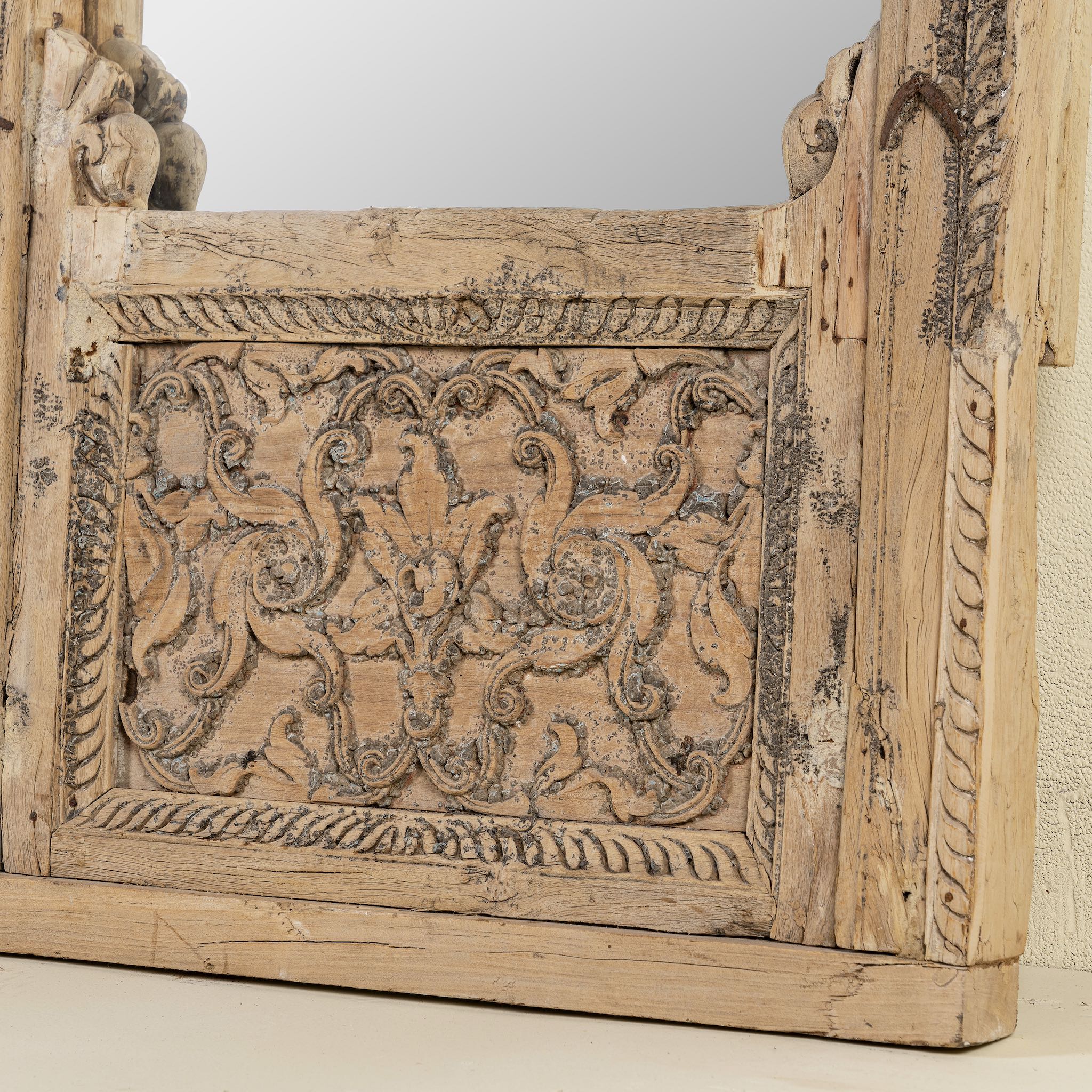 Close up of the base of a hand-carved mirror crafted from an old teak wood window frame, featuring intricate floral and scrollwork details with a natural distressed finish and an arched top design reflecting traditional Indian craftsmanship.