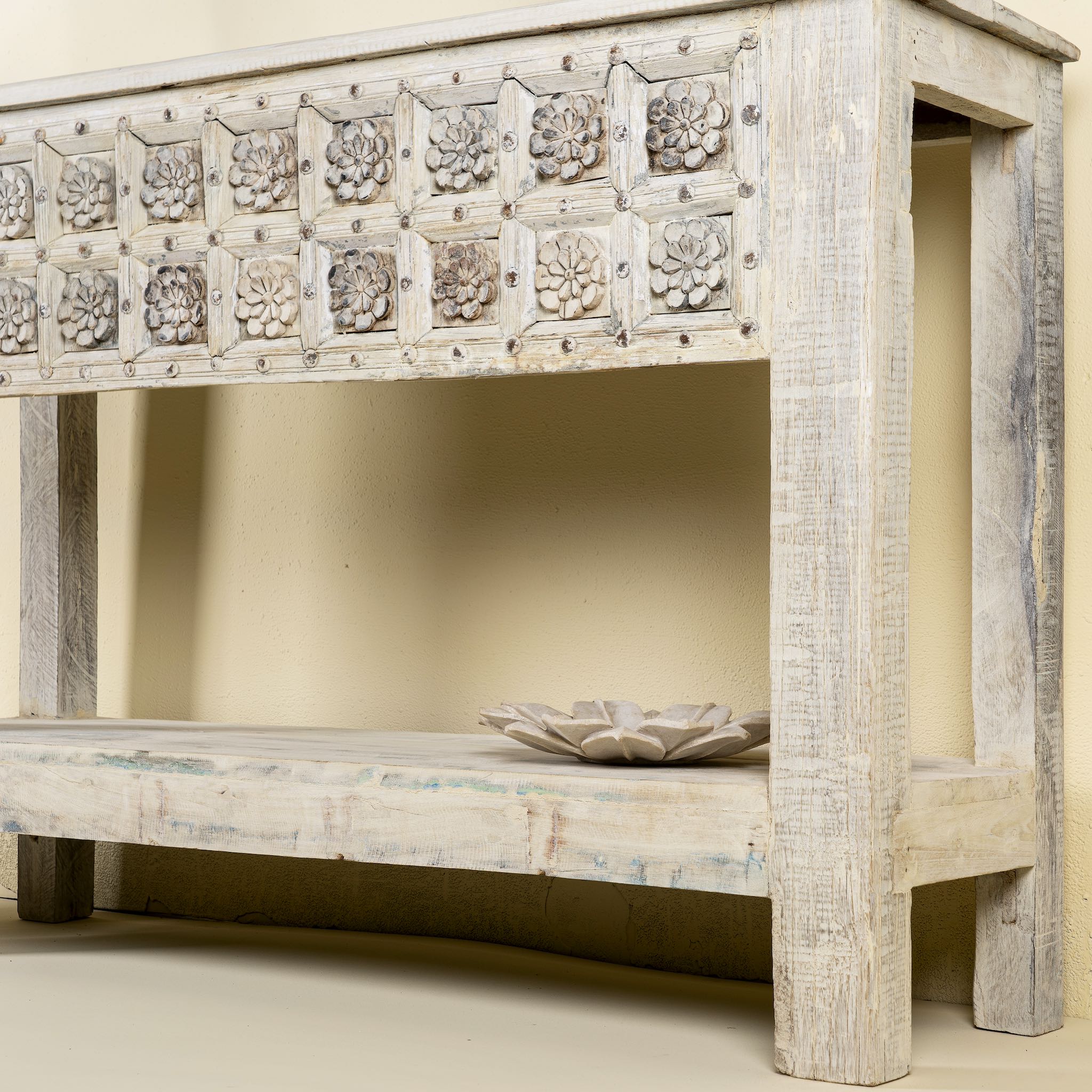 Close up side angle of a handcarved console table made from reclaimed teak wood, featuring a detailed front panel with geometric floral motifs and a distressed whitewashed finish, complemented by a lower shelf for added storage or display.