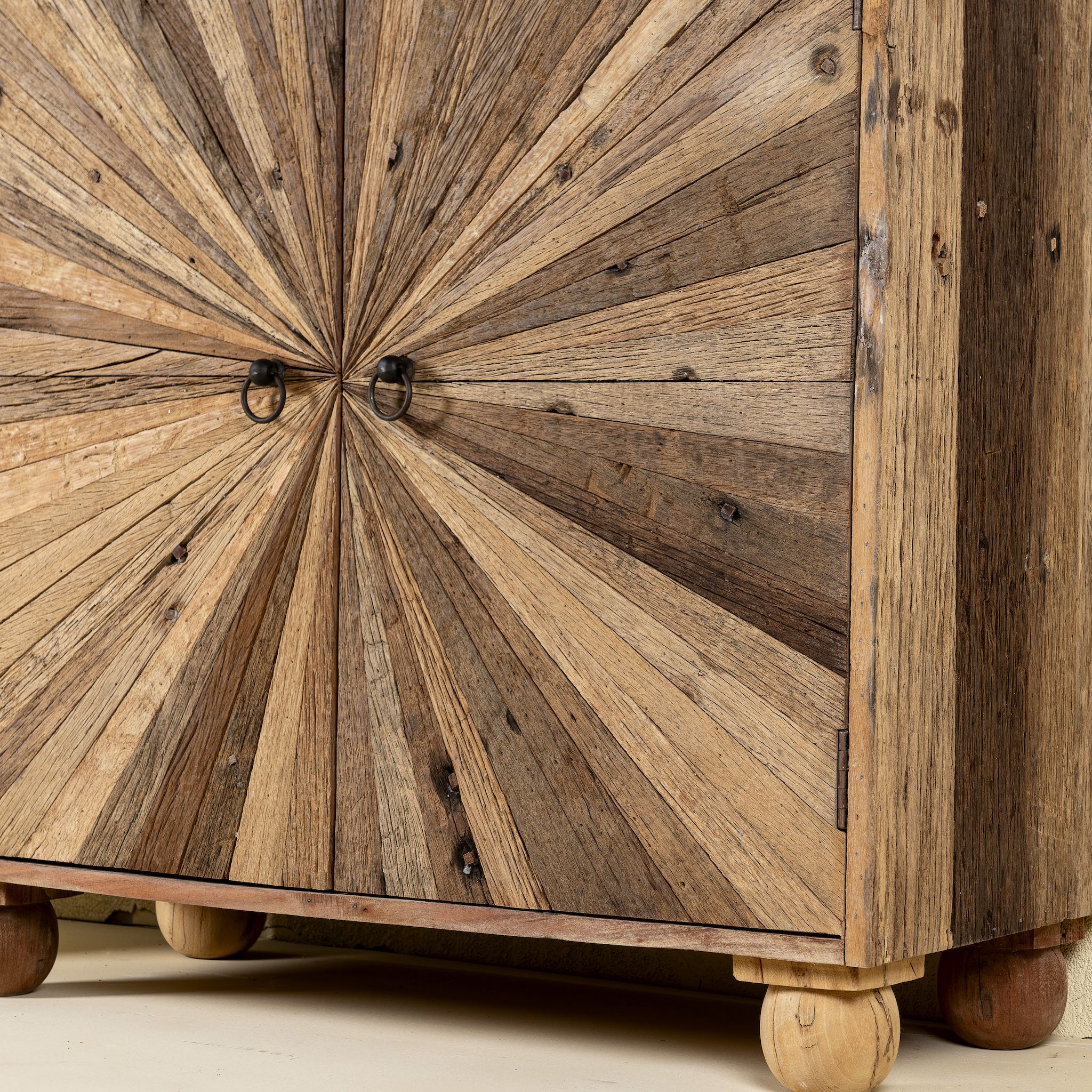 Close up leg image of a artistic wooden cabinet in rustic style, made from reclaimed wood with a striking sunburst pattern on the double doors, featuring subtle color variations and round metal ring pulls, elevated on rounded wooden feet.