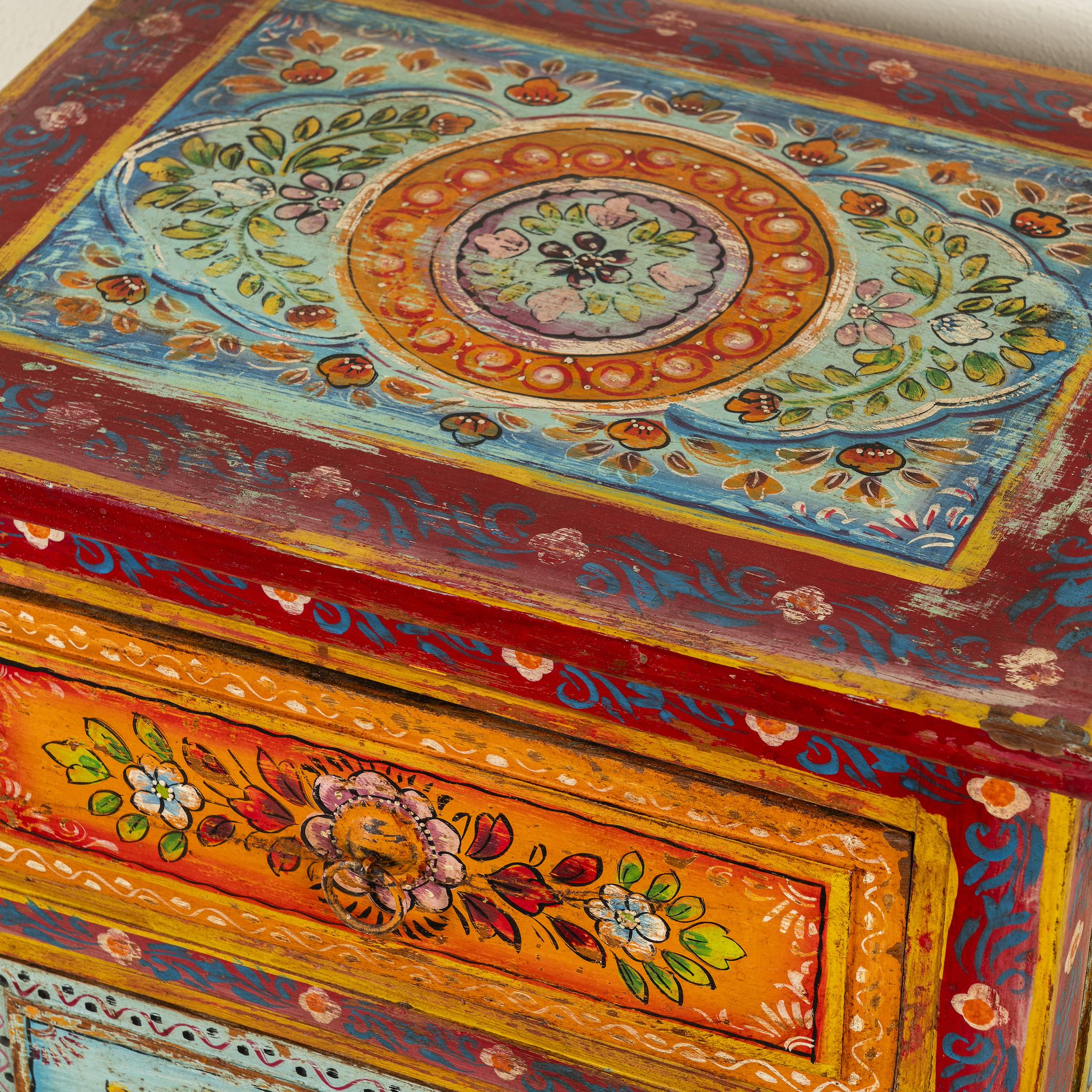 Close up image of a hand-painted reclaimed wood nightstand with multiple drawers, featuring vibrant colours and floral patterns, placed next to a vase with dried plants.