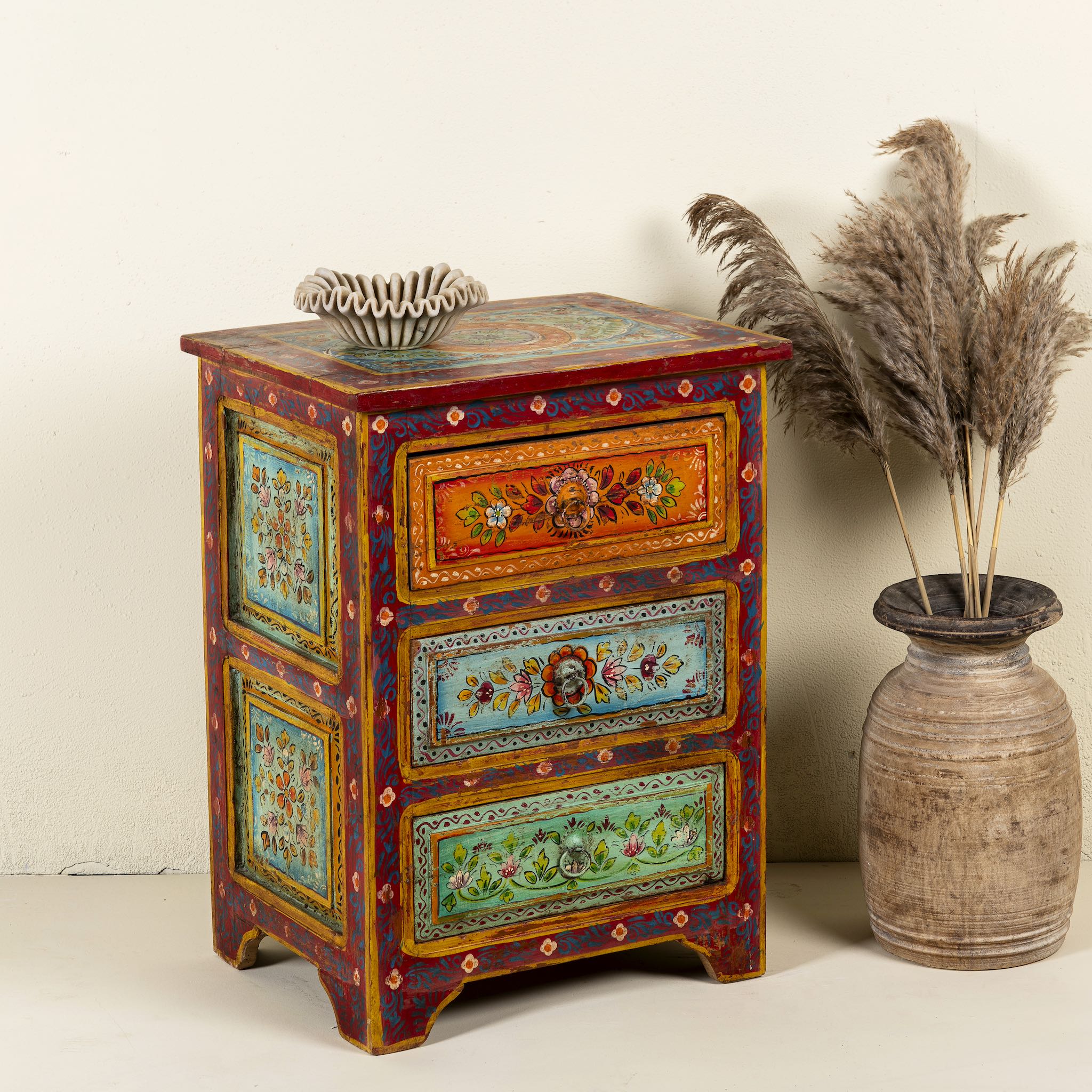 Side angle image of a hand-painted reclaimed wood nightstand with multiple drawers, featuring vibrant colours and floral patterns, placed next to a vase with dried plants.