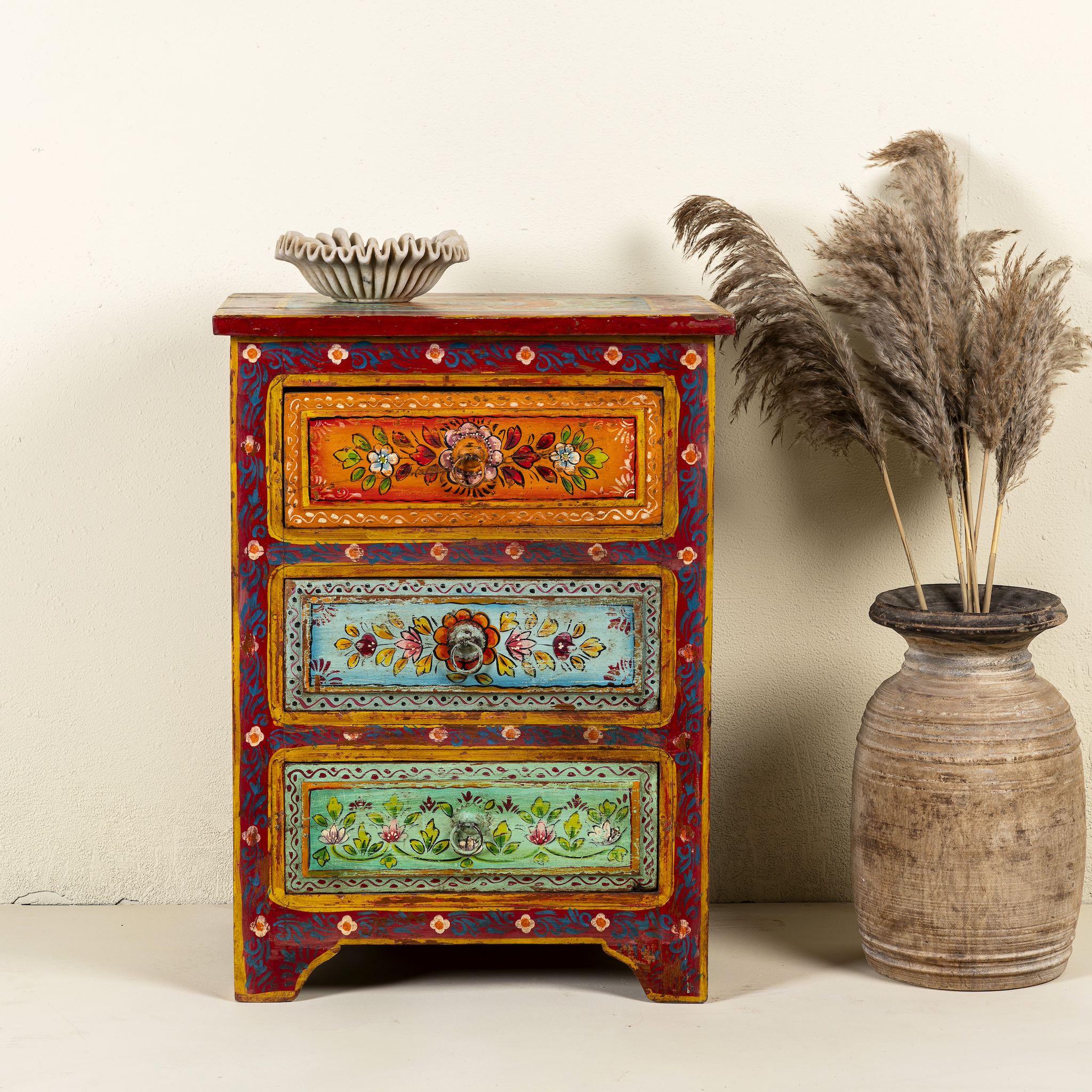 A hand-painted reclaimed wood nightstand with multiple drawers, featuring vibrant colours and floral patterns, placed next to a vase with dried plants.