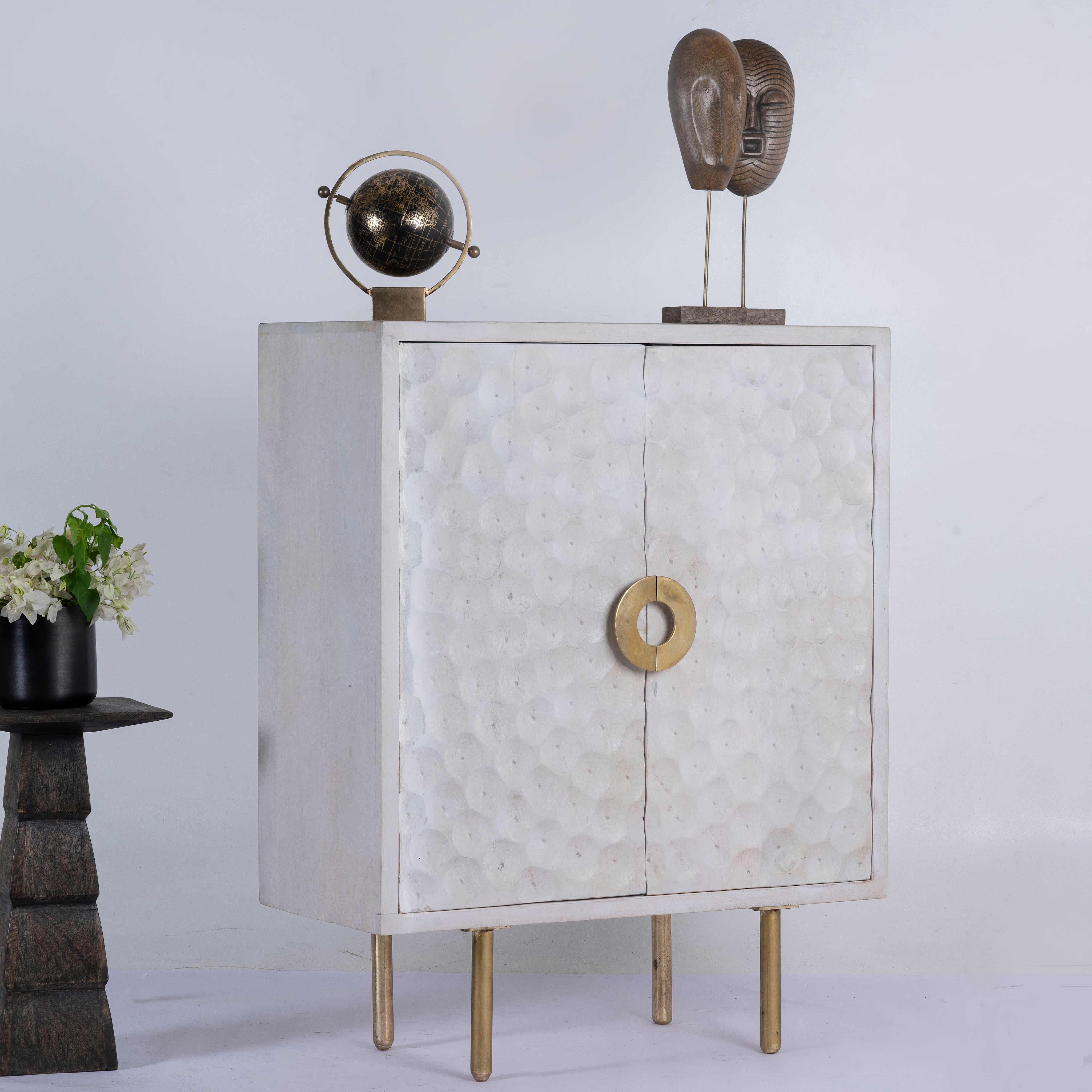 Side angle image of a carved wood furniture cupboard in white mango wood with hammered pattern, brass iron base, 88 × 43 × 122 cm.