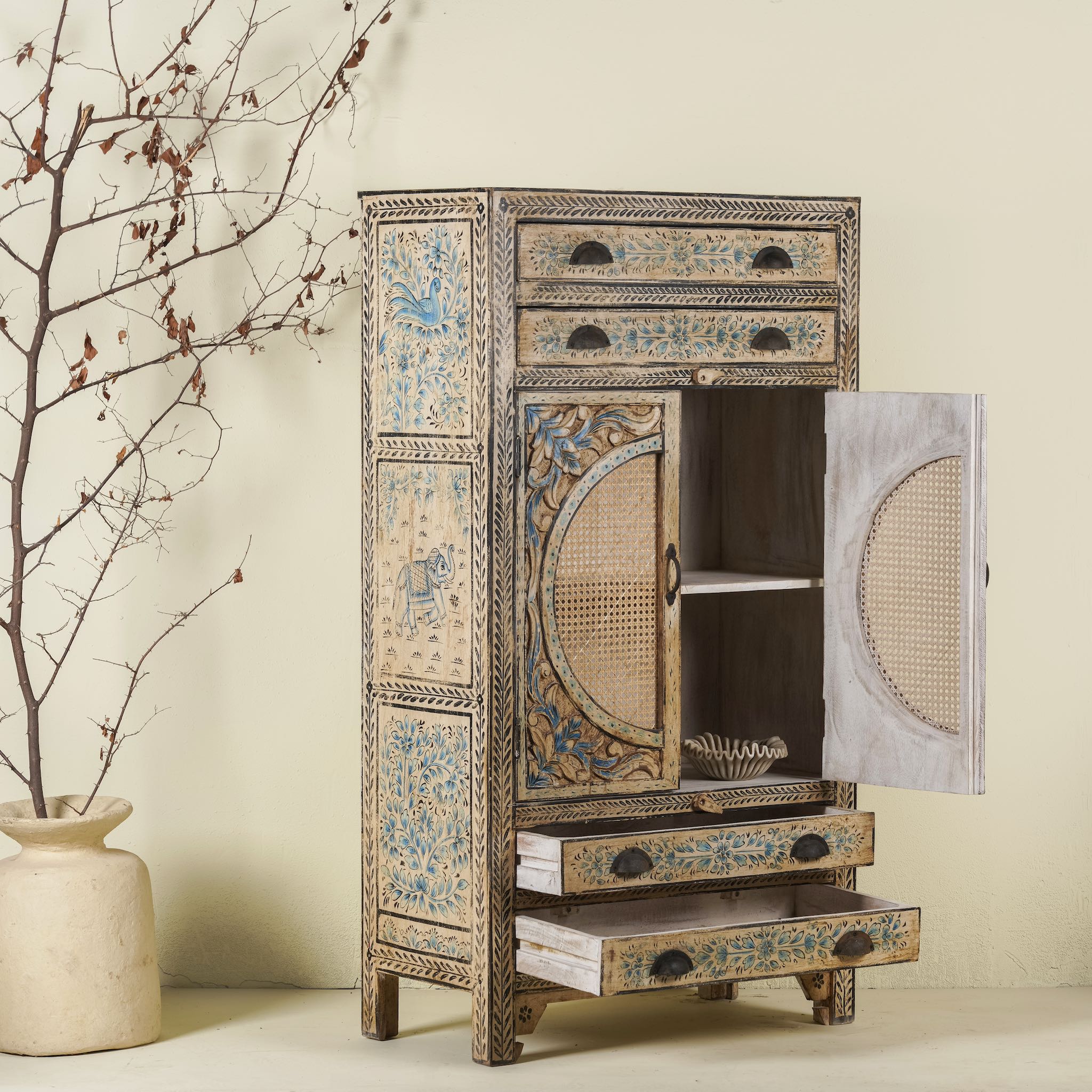 Open door image of a hand-painted cabinet made from mango wood, featuring rattan inlay doors framed by a carved circular motif, floral blue and green accents, and five drawers for additional storage.
