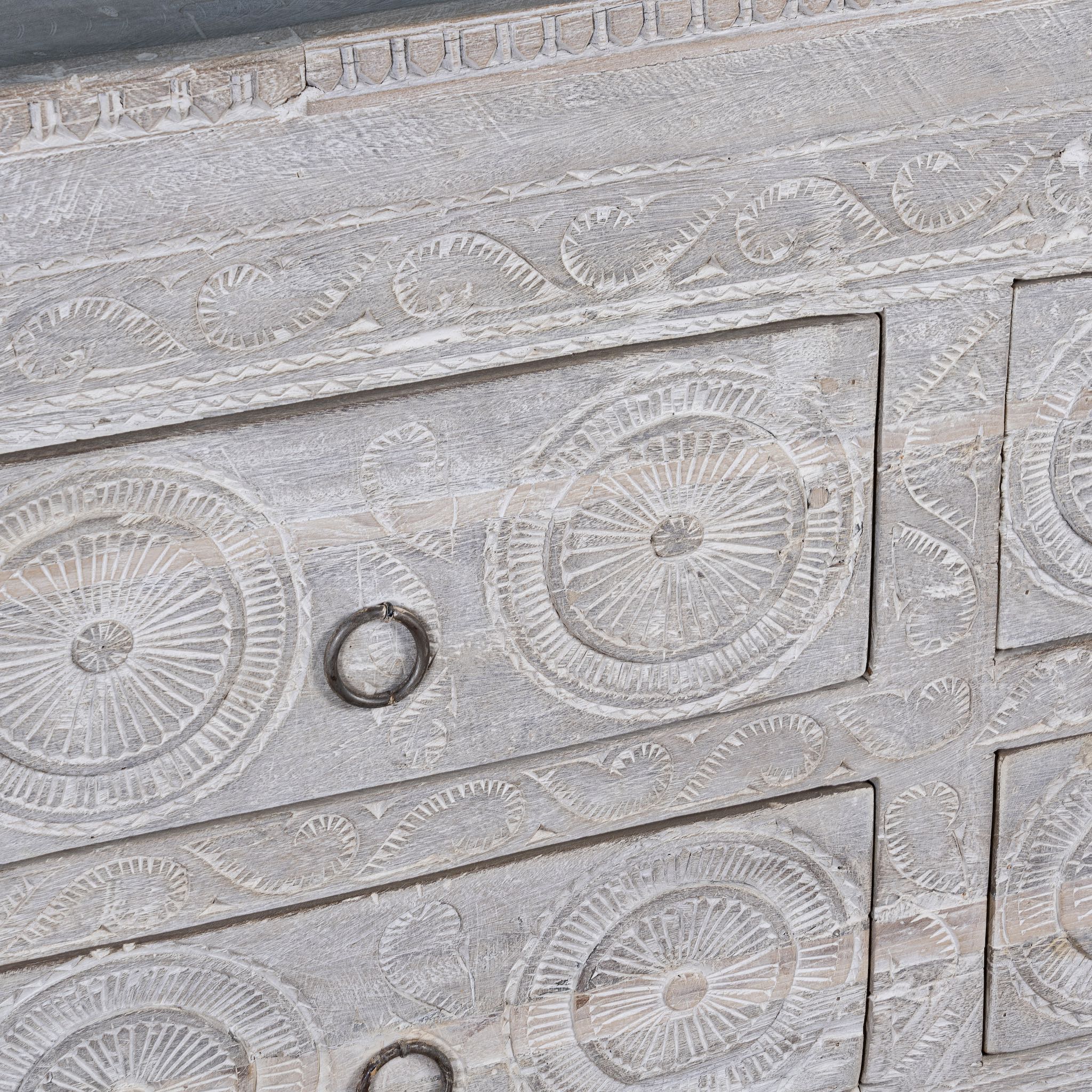 Close up view of a Ibiza style sideboard with six drawers, made from hand-carved wood in a whitewashed finish, featuring circular mandala motifs and rustic iron handles.