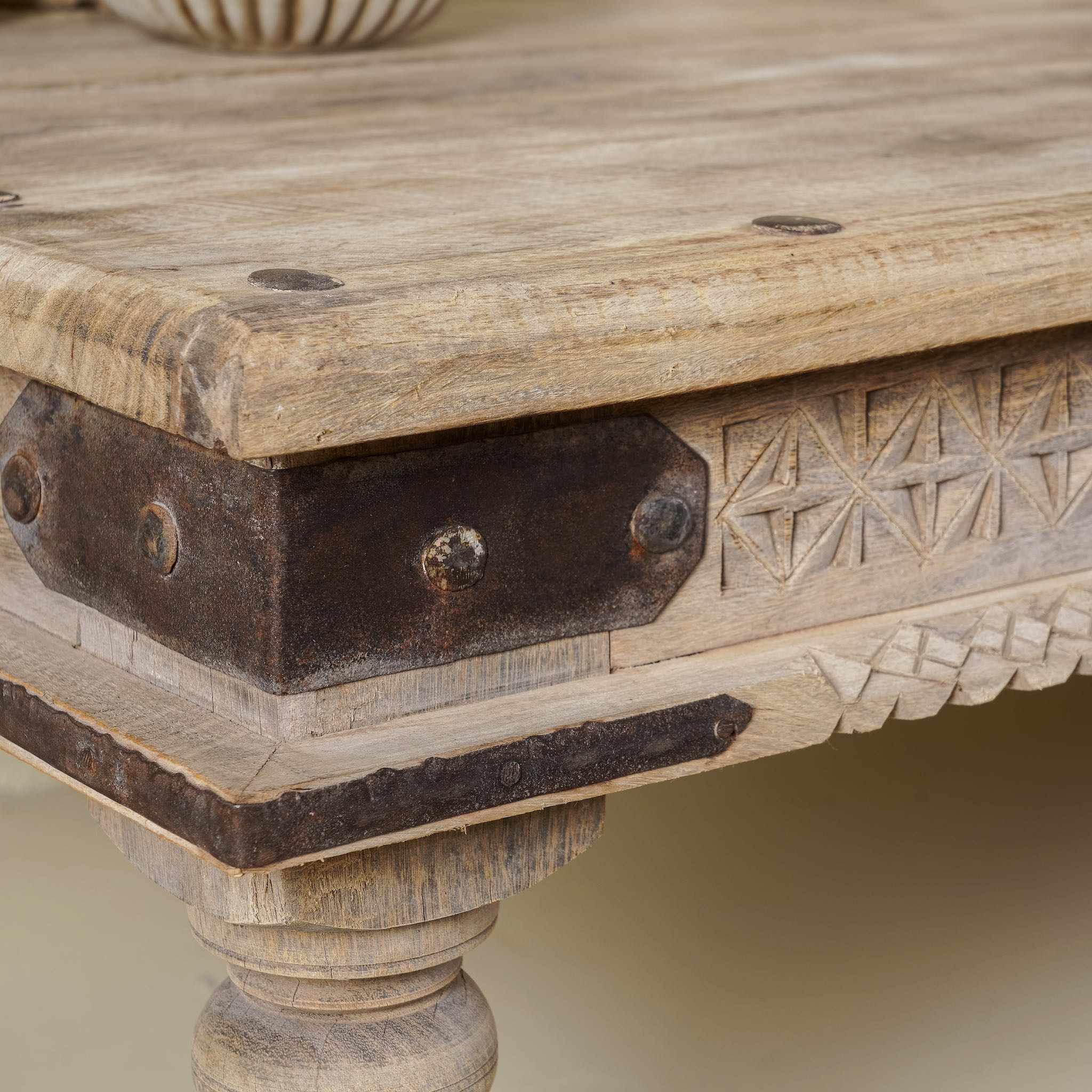 Close up of a corner of a rustic coffee table made from old reclaimed teak wood, featuring a weathered finish, turned legs, decorative carved apron, and visible metal rivets, styled with earthy decor in a minimal interior setting.