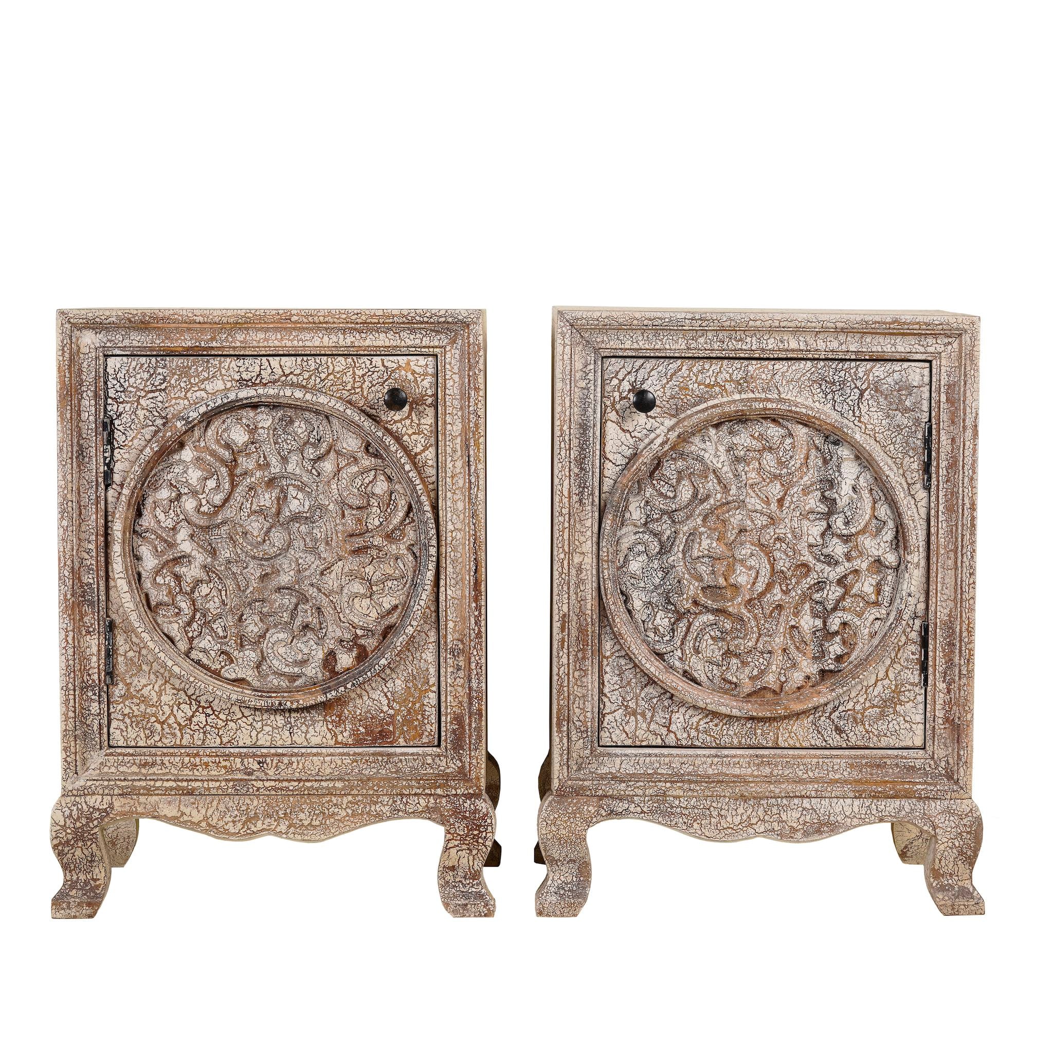 White background picture of a pair of hand-carved bedside tables made of mango wood, featuring intricate round floral motifs on the doors, crackled vintage finish, and elegant curved legs in a distressed antique design.