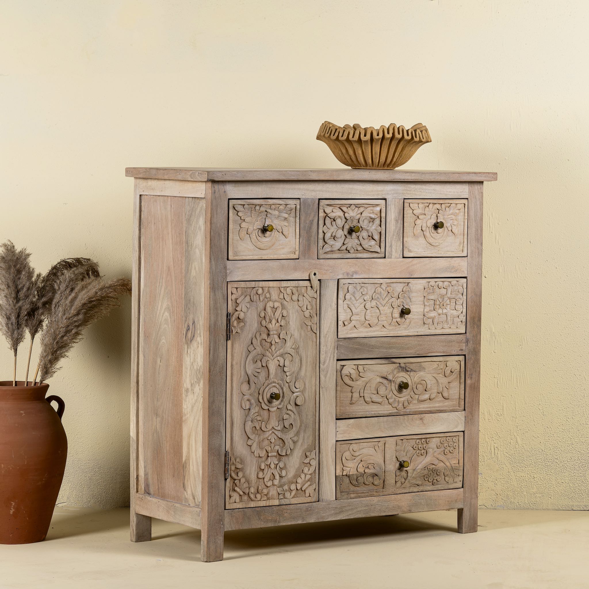 Side view of a hand carved mango wood cupboard with 6 drawers and 1 door, iron fitted door knobs and floral carvings.