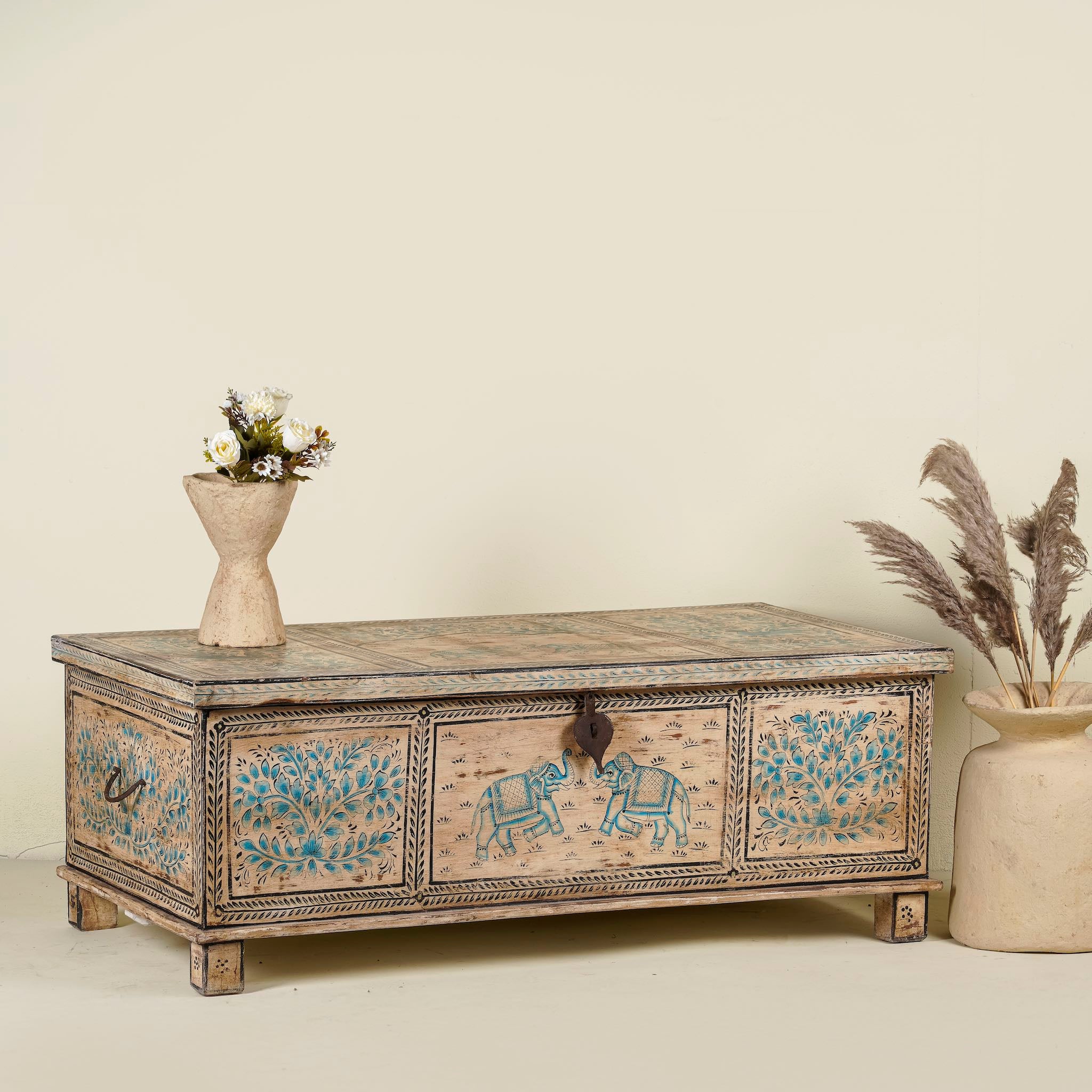 Side angle image of a large hand-painted chest made from old reclaimed wood, featuring intricate blue designs of elephants and floral motifs on a distressed beige background, with a central latch and a rustic vintage finish.