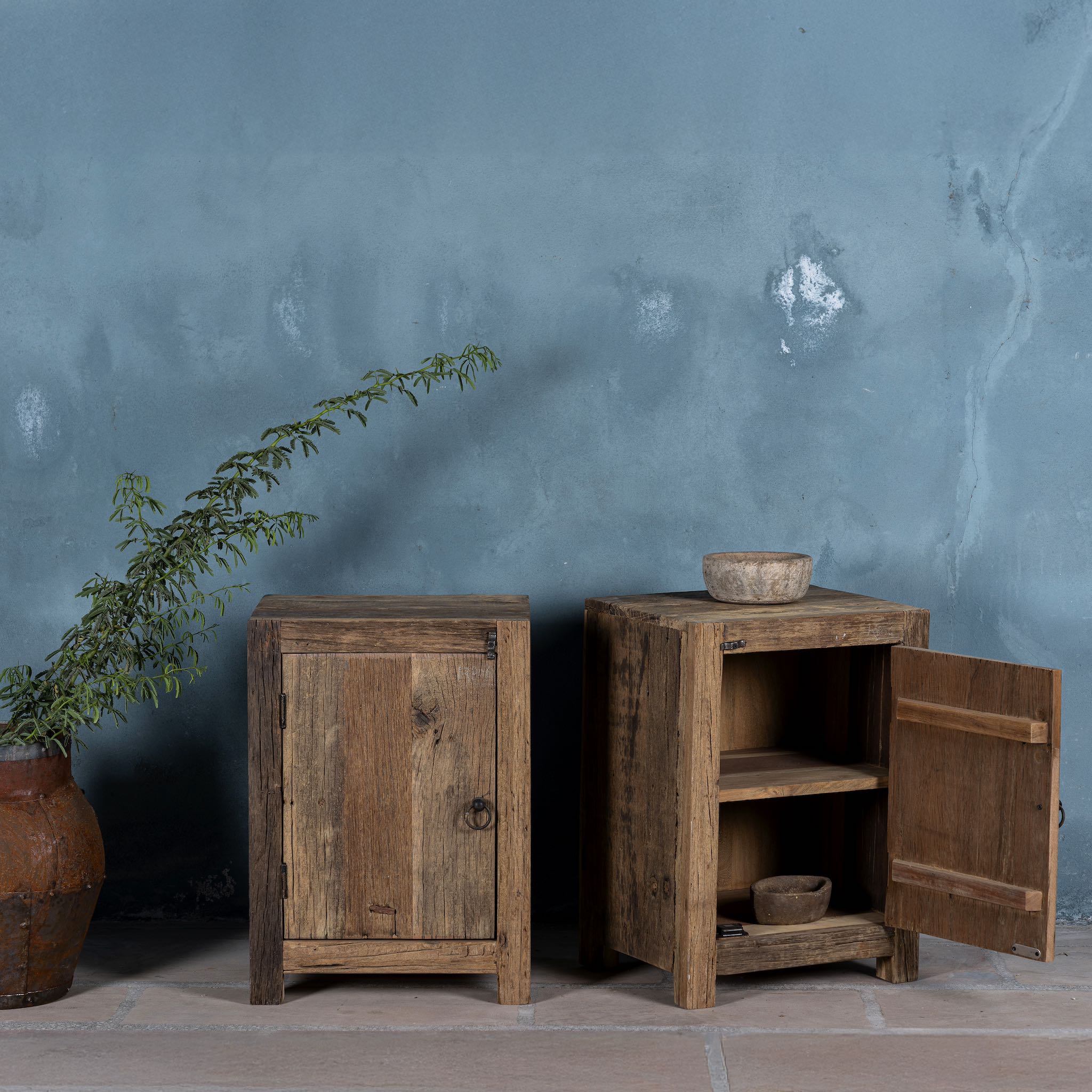 Open door view of a pair of night stands in rustic minimalistic design with iron door handles decorated in front of a blue wall.