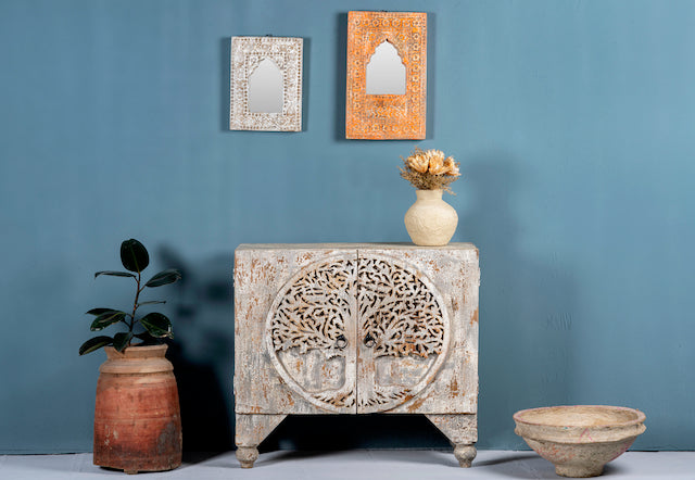 A beige mango wood cupboard with intricate hand-carved tree designs on the front, with a framed mirror above, a potted plant to the left, and a decorative bowl to the right on a blue background.