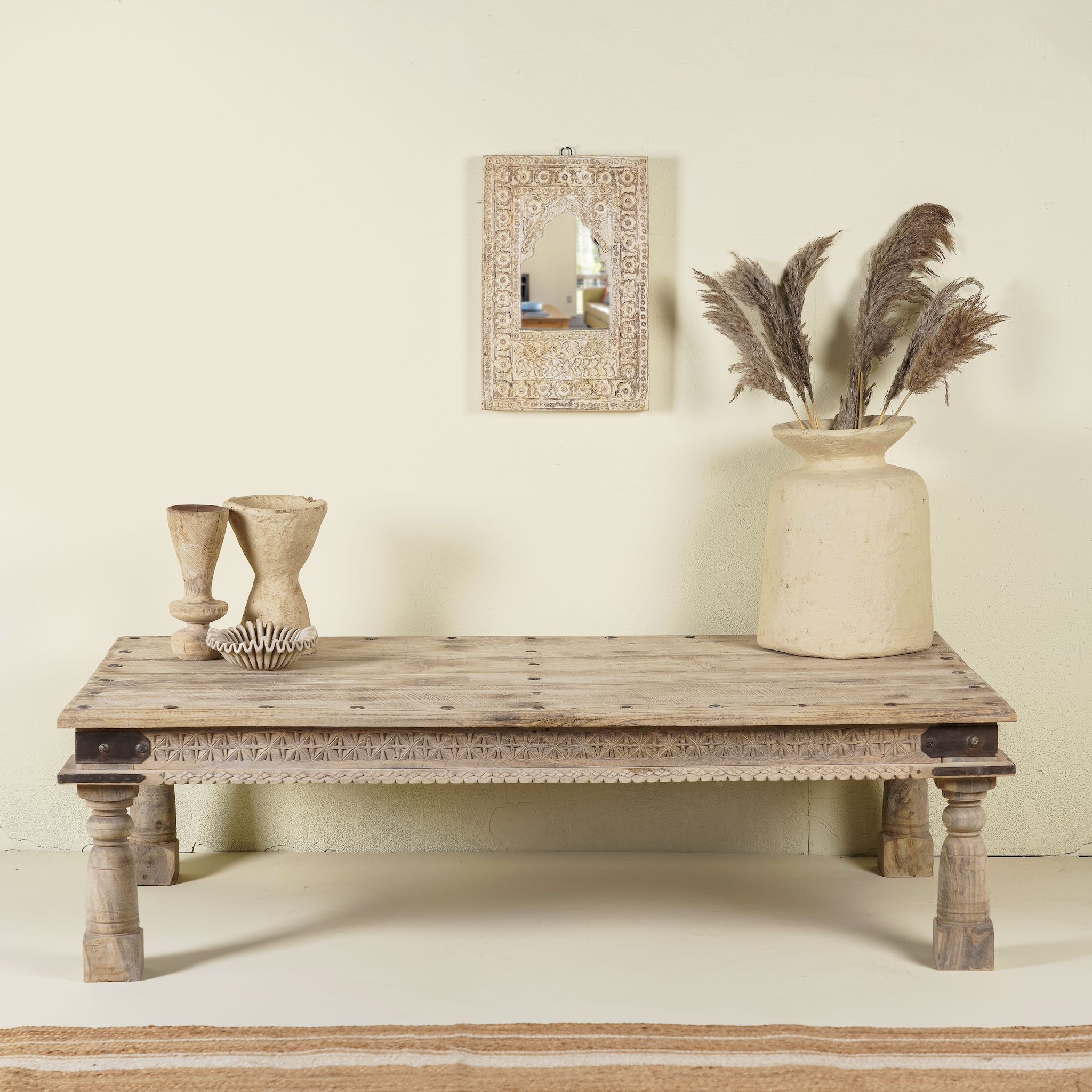 Rustic coffee table Ritvik made from old reclaimed teak wood, featuring a weathered finish, turned legs, decorative carved apron, and visible metal rivets, styled with earthy decor in a minimal interior setting.