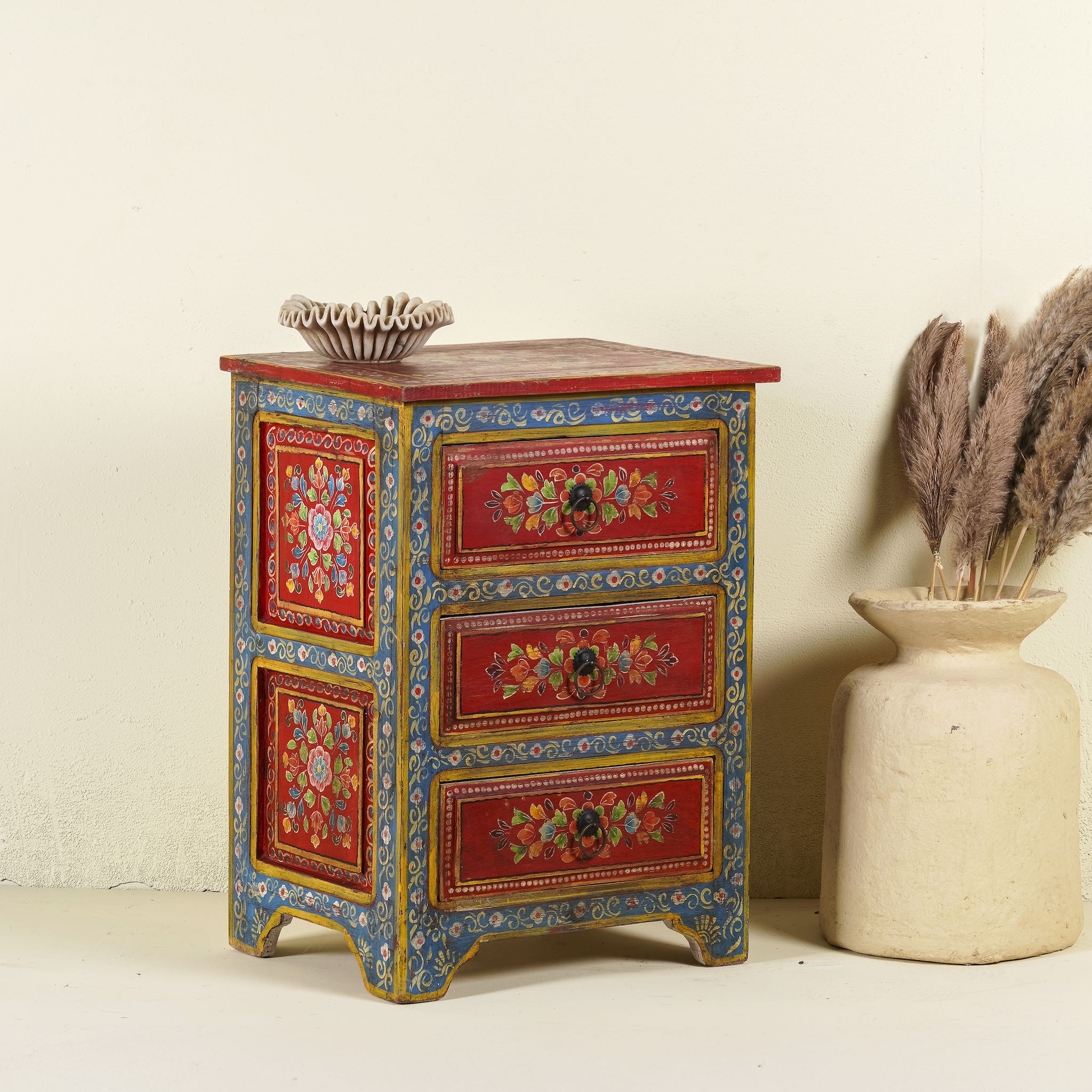 Side angle image of a hand-painted reclaimed wood nightstand with vibrant floral motifs, featuring three drawers and a recycled wood top, placed next to a vase with dried plants.