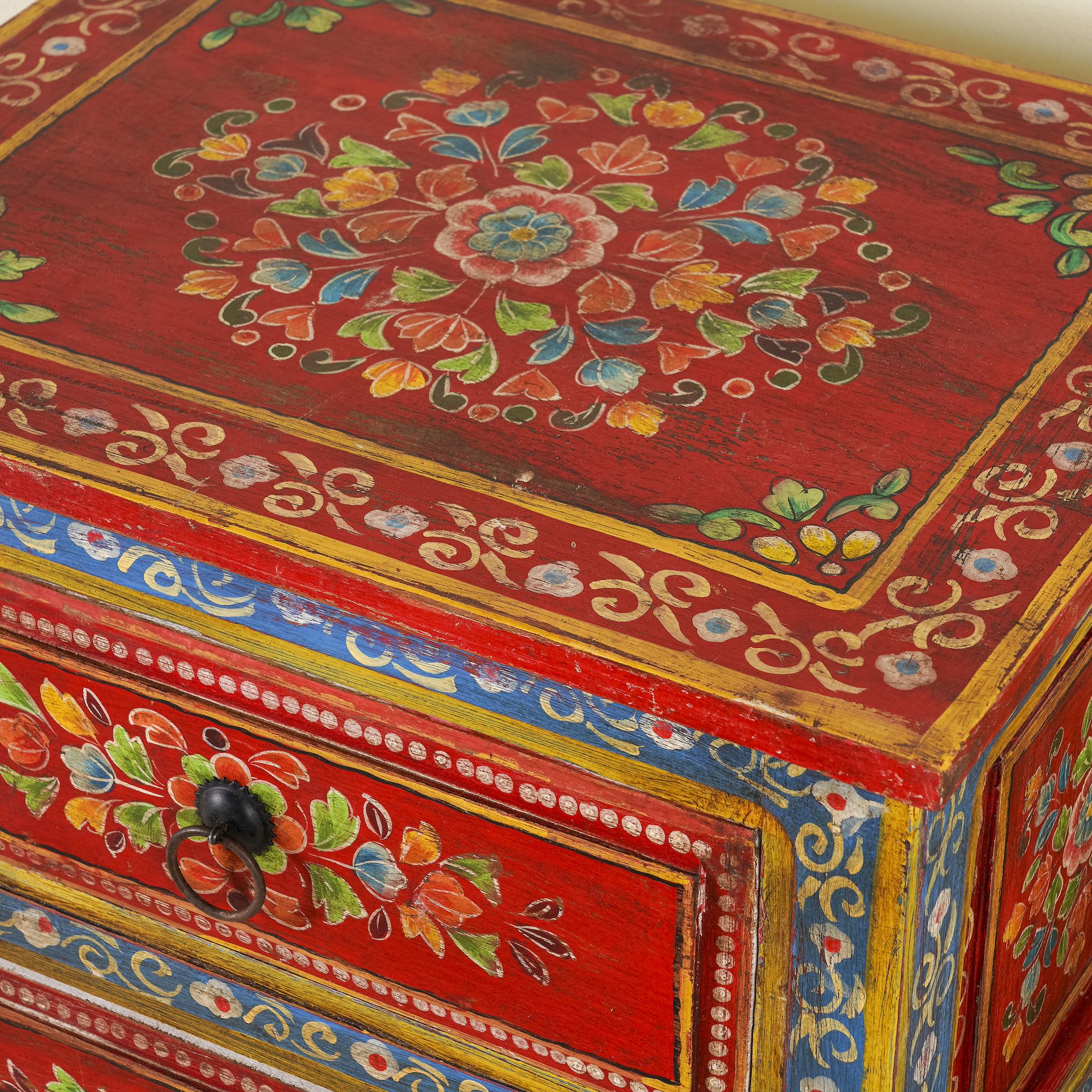 Close up image of a hand-painted reclaimed wood nightstand with vibrant floral motifs, featuring three drawers and a recycled wood top, placed next to a vase with dried plants.