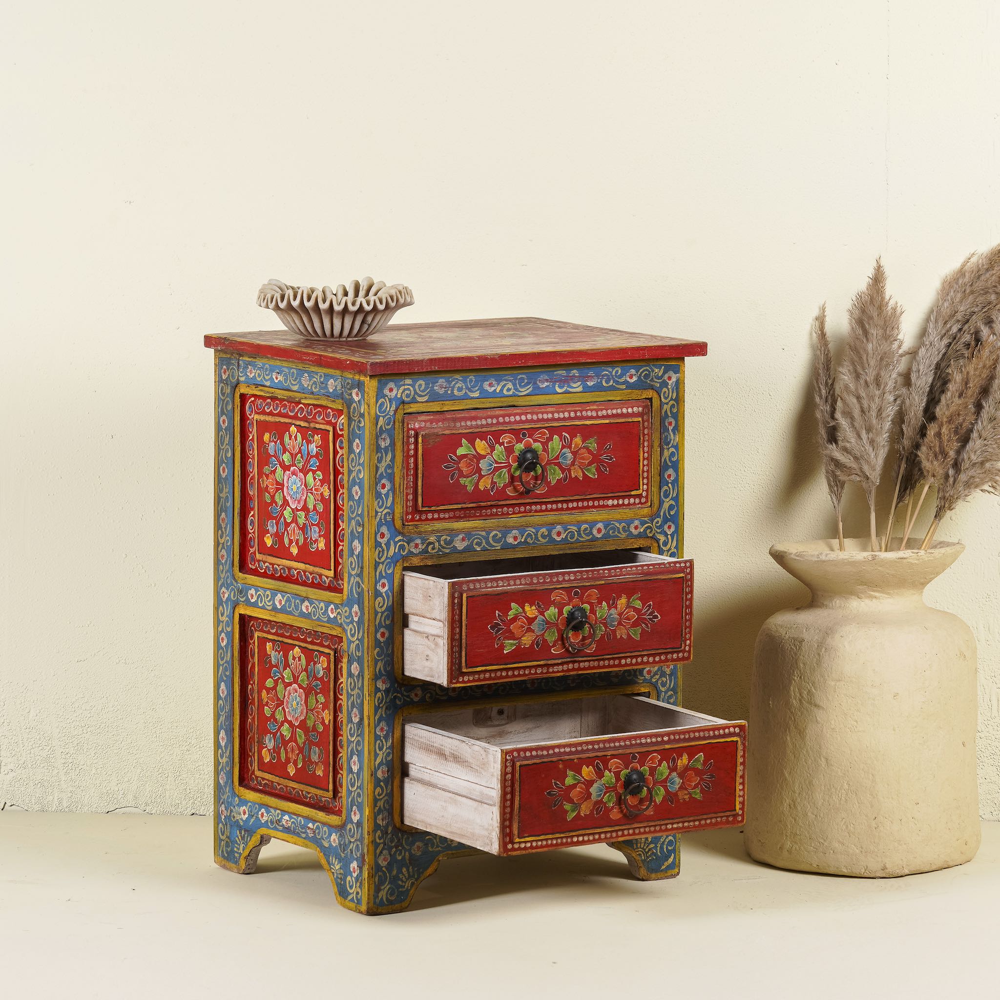 Open drawer image of a hand-painted reclaimed wood nightstand with vibrant floral motifs, featuring three drawers and a recycled wood top, placed next to a vase with dried plants.