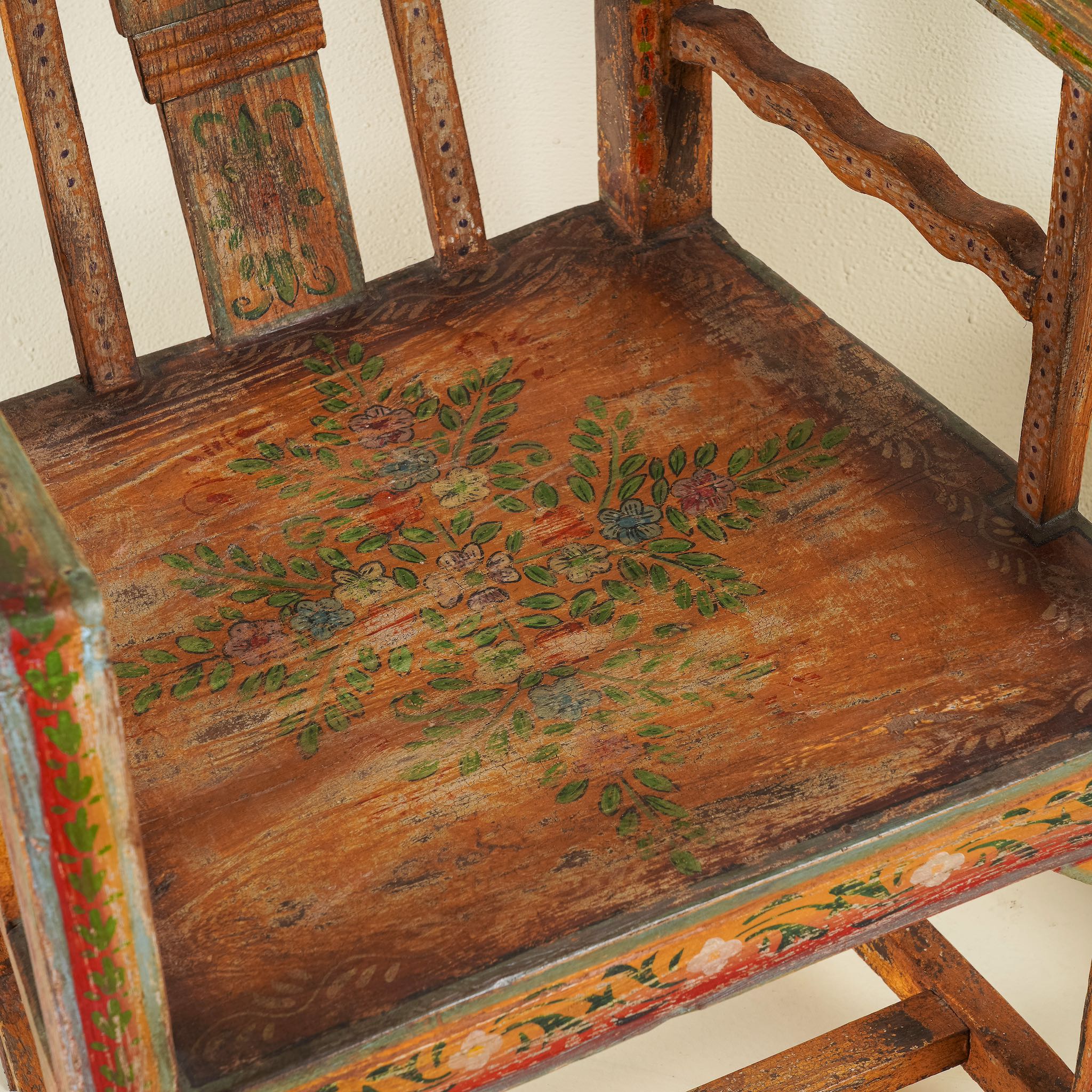 Close up image of a hand-painted armchair crafted from reclaimed teak wood, featuring intricate traditional patterns in earthy tones on the backrest, seat, and frame, with a rustic and vintage finish.