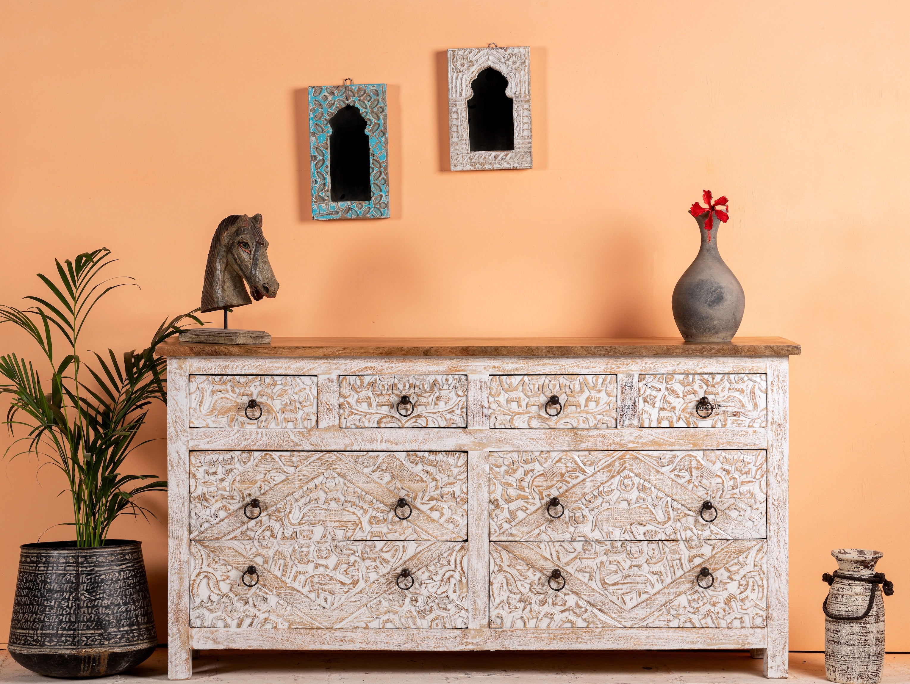 A hand-carved mango wood sideboard with whitewashed finish featuring elephant motifs. The sideboard is placed in a room with a plant, decorative items, and wall hangings.