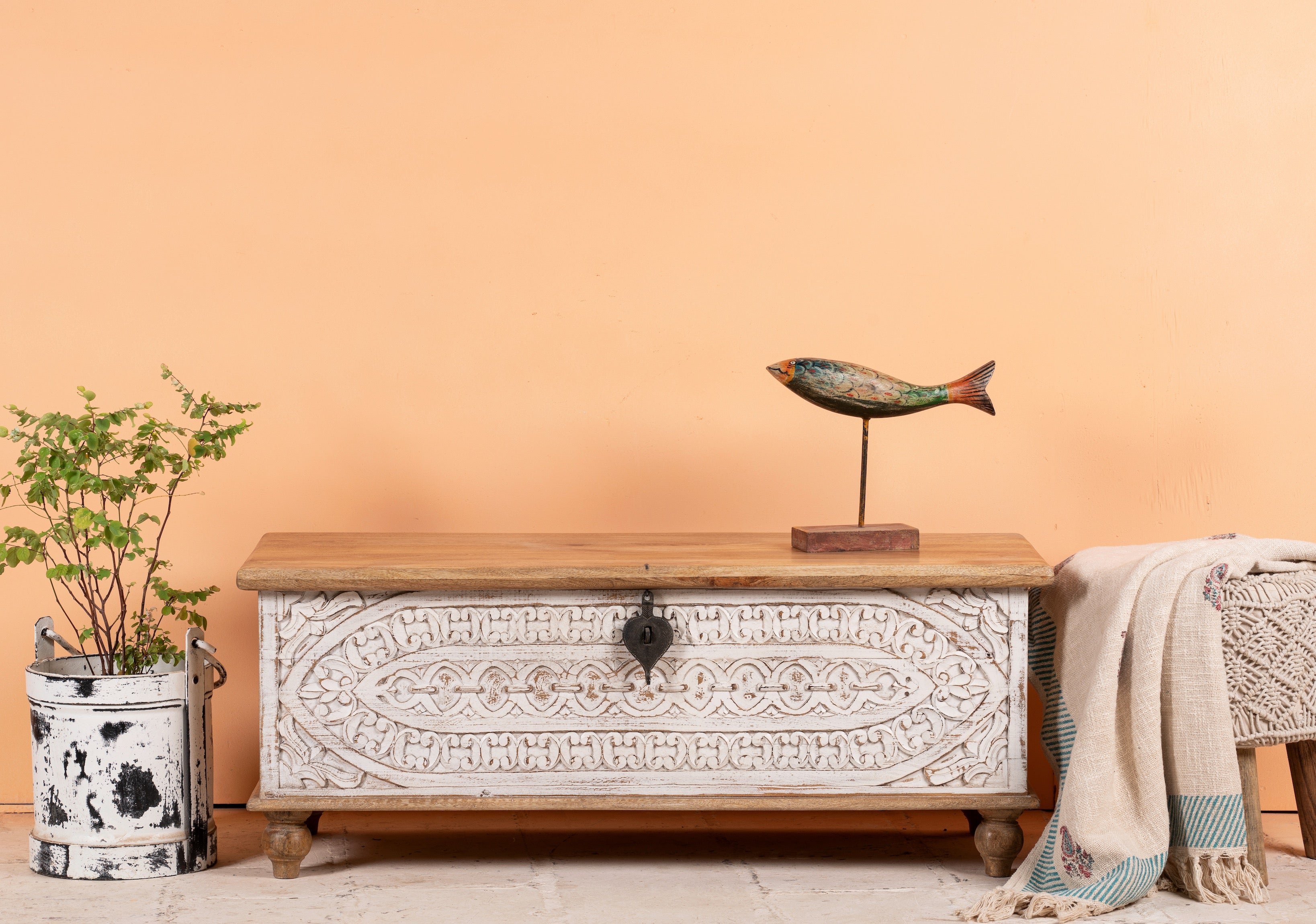 A hand-carved mango wood storage box with intricate patterns and antique-style iron fittings, finished in a distressed white, placed in a room with a plant and a decorative fish figure on top.