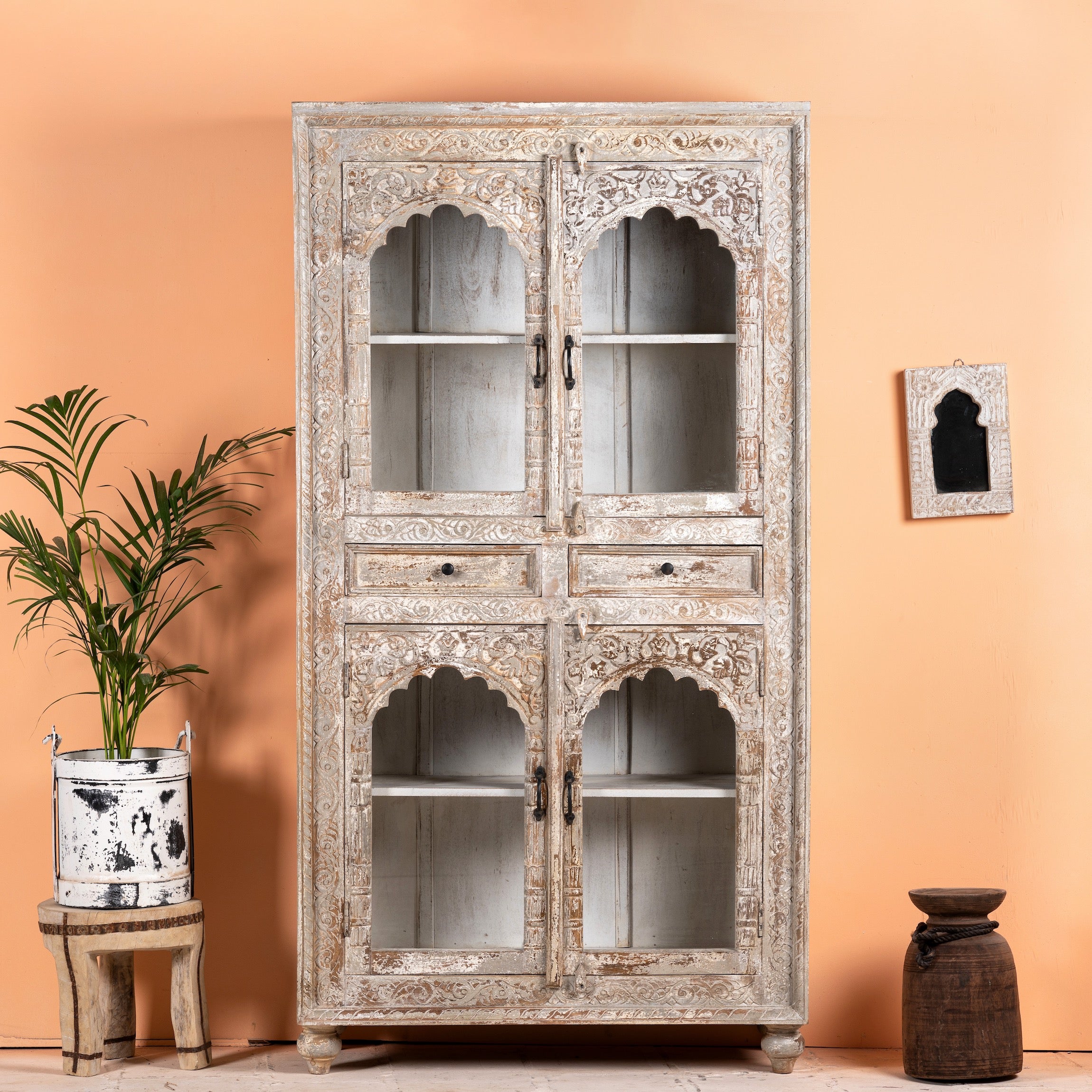 An oriental-style Showcase Palitana cabinet with four glass doors and hand-carved Indian patterns on the front, displayed in a home setting with furniture and decorative items around it.