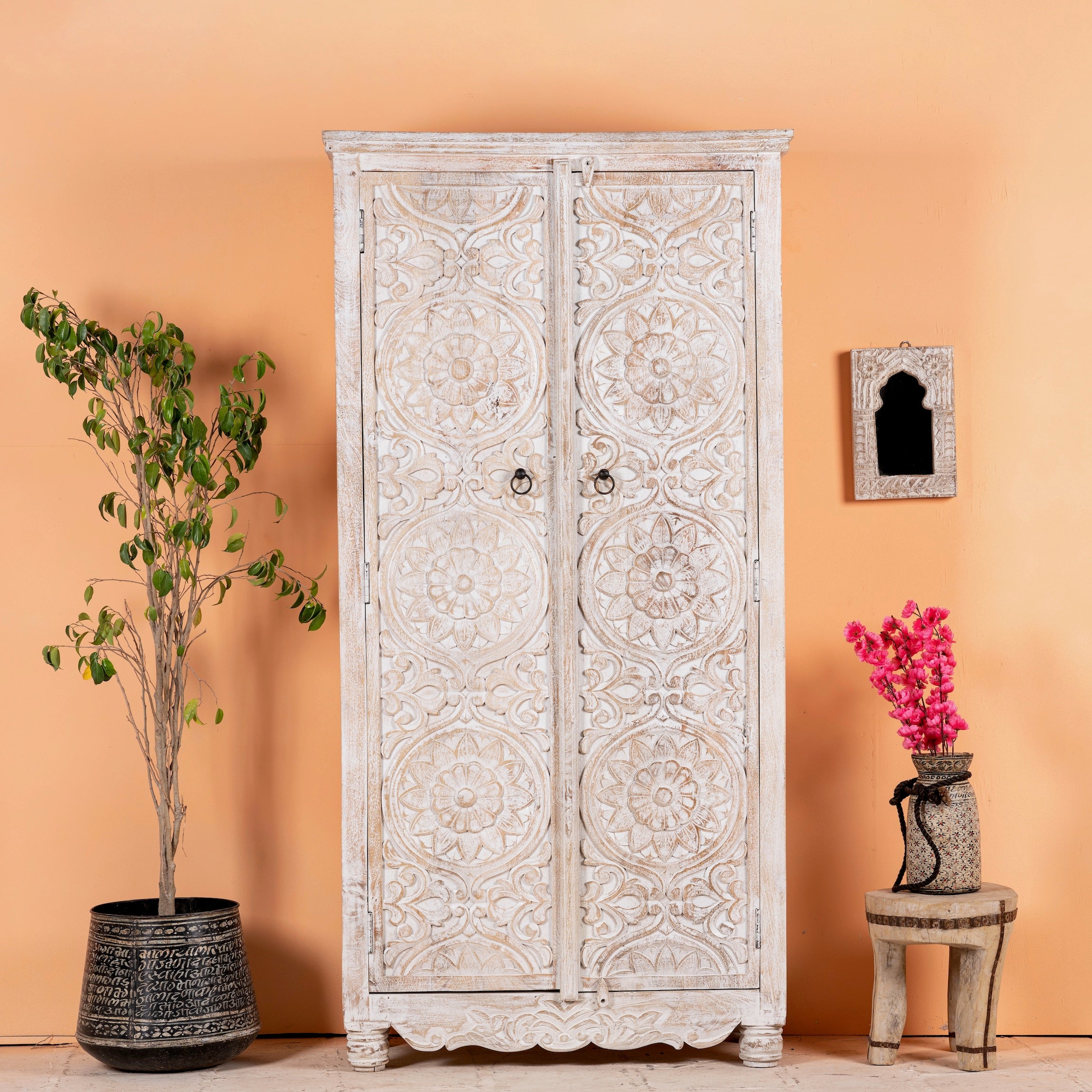 A white-washed, hand-carved mango wood cabinet with intricate patterns, placed against an orange wall next to a potted plant and a decorative object.