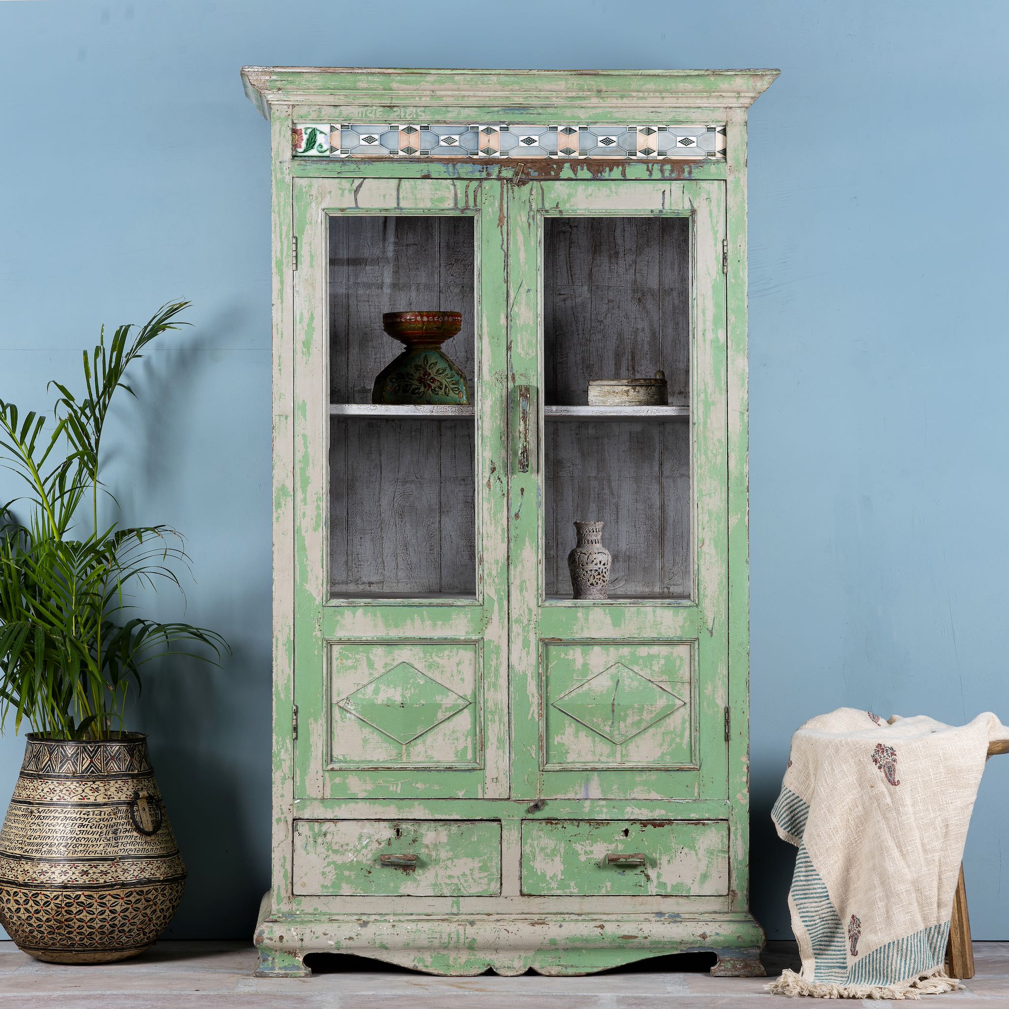 Reclaimed teak wood vitrine with distressed green finish, ceramic inlays, and glass-panelled doors – sustainable furniture with vintage Indian charm.