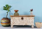A hand-carved mango wood storage box with floral patterns, a whitewashed finish, and iron handles, displayed against a blue wall with vase and flower arrangement.