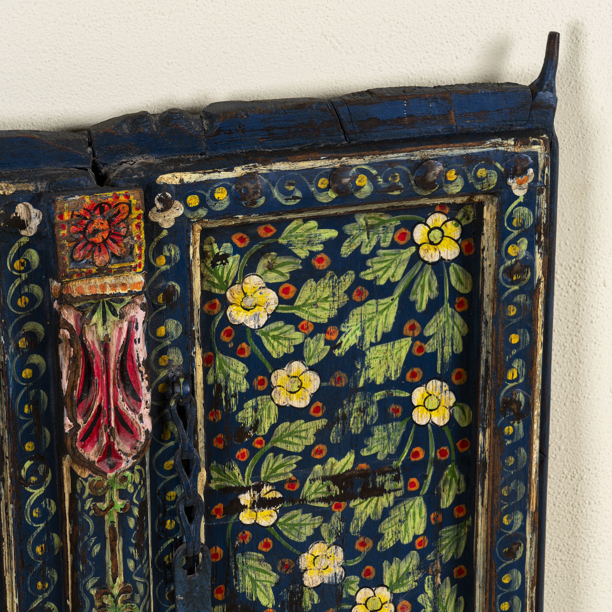 Close up image of a decorative wall panel made from an old teak wood window frame, hand-painted with intricate floral patterns in vibrant colours including green, red, yellow, and blue, showcasing traditional Indian craftsmanship and vintage charm.
