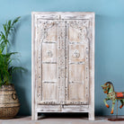 A white-washed, wooden cabinet with intricate iron applications, standing in a room with a potted plant and a decorative horse figure.