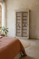 Lifestyle image of a old door cabinet with reclaimed teak doors, floral carvings, and iron accents; artisan teak wood wardrobe with mango wood frame, 100 × 44 × 206 cm.