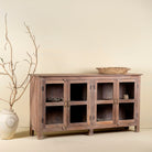 Side angle image of a rustic showcase cabinet made from recycled teak wood, featuring four glass-paneled doors with brass handles and a lightly weathered finish, offering a warm, natural look ideal for displaying decor or tableware.
