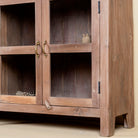 Close up image of the leg of a rustic showcase cabinet made from recycled teak wood, featuring four glass-paneled doors with brass handles and a lightly weathered finish, offering a warm, natural look ideal for displaying decor or tableware.
