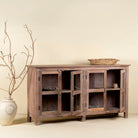 Side angle with open doors image of a rustic showcase cabinet made from recycled teak wood, featuring four glass-paneled doors with brass handles and a lightly weathered finish, offering a warm, natural look ideal for displaying decor or tableware.
