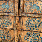 Close up image of a hand-painted wooden wall panel made from an old window frame, featuring intricate blue floral patterns along with stylised animal motifs such as elephants and camels, set against a distressed golden-brown background with an arched top frame.
