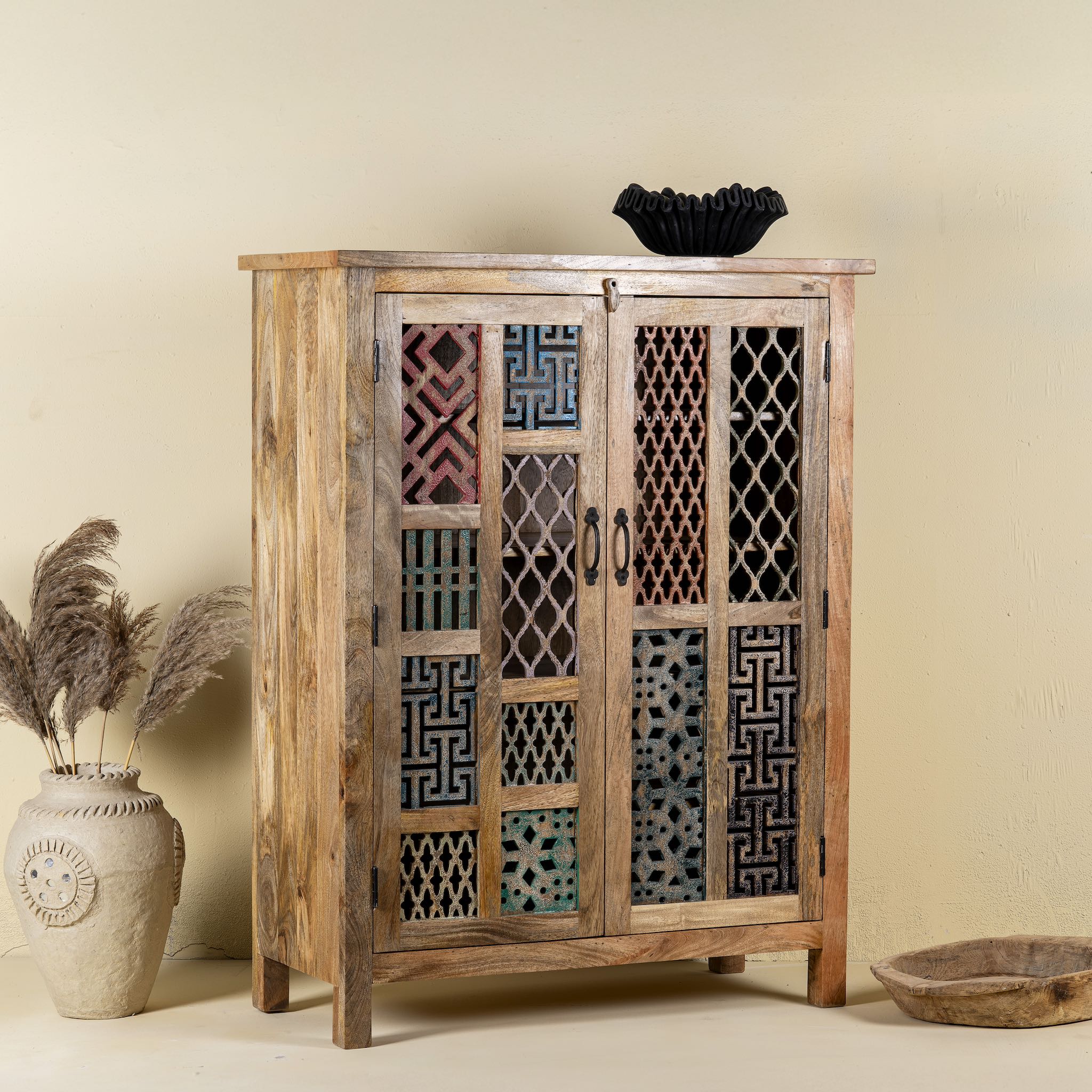 Side angle image of a artisan-made cabinet crafted from mango wood, featuring double doors adorned with a patchwork of colourful, intricately carved geometric panels, blending traditional craftsmanship with a vibrant, eclectic style.
