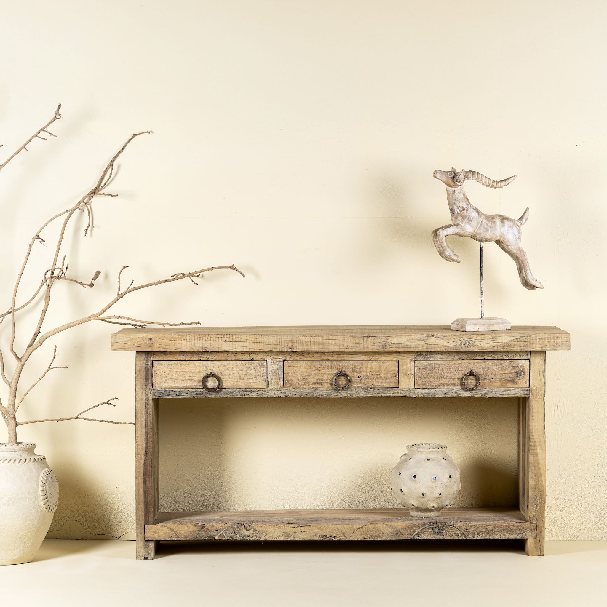Front view image of a rustic console table crafted from reclaimed teak wood, featuring a natural weathered finish, three drawers with metal ring pulls, and a lower shelf for additional storage or display, blending simplicity with organic charm.
