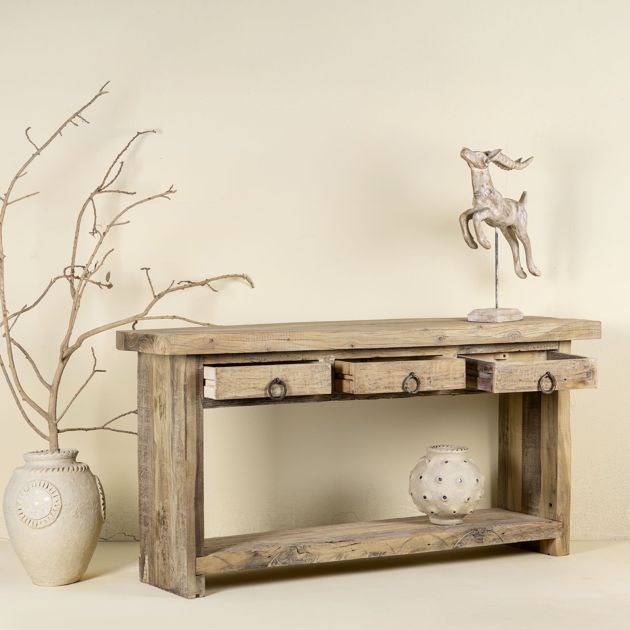 Side angle image with open drawers of a rustic console table crafted from reclaimed teak wood, featuring a natural weathered finish, three drawers with metal ring pulls, and a lower shelf for additional storage or display, blending simplicity with organic charm.
